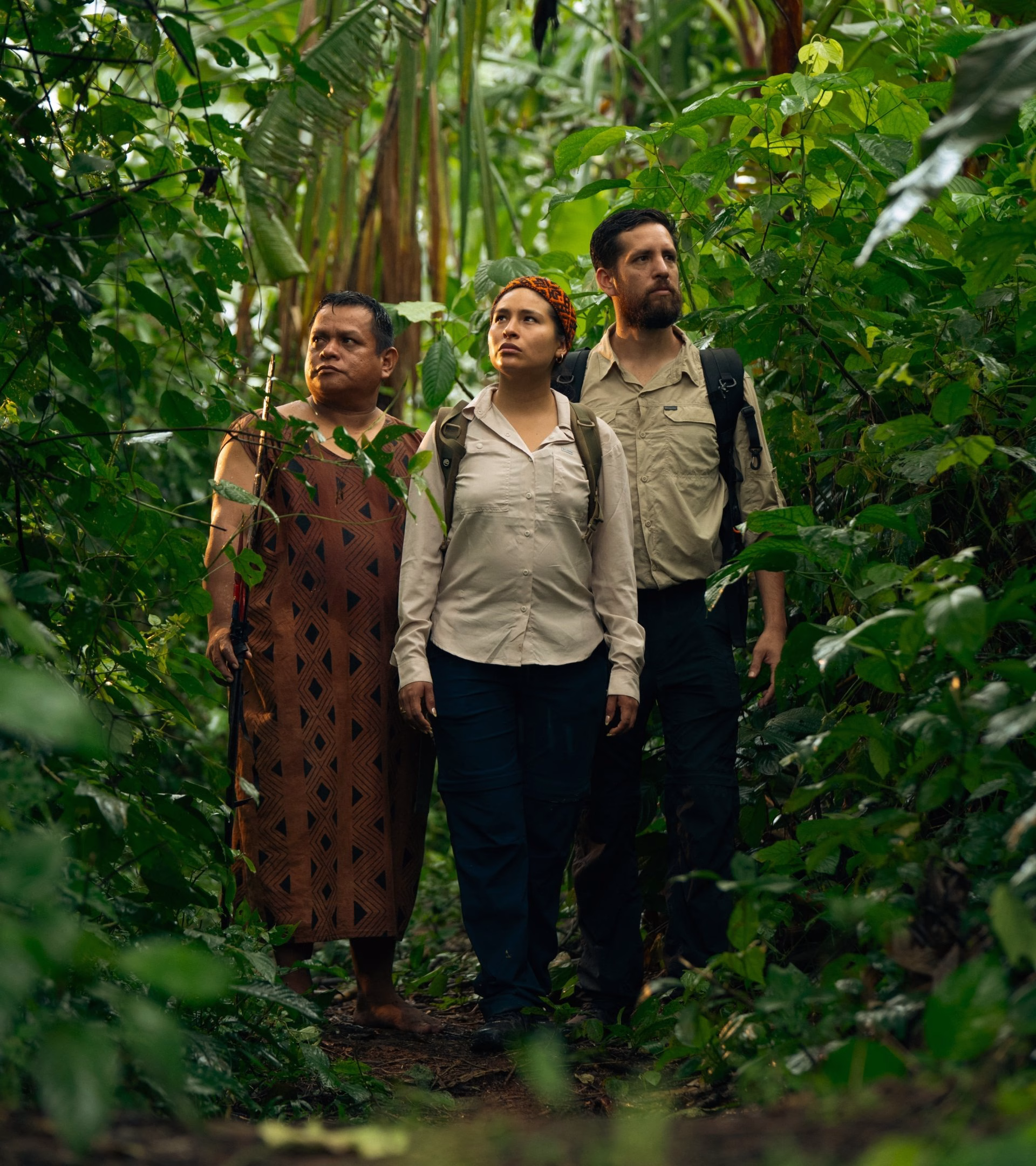 Three individuals stand in a lush green jungle, embodying indigenous narratives with modern attire and expressions of contemplation.