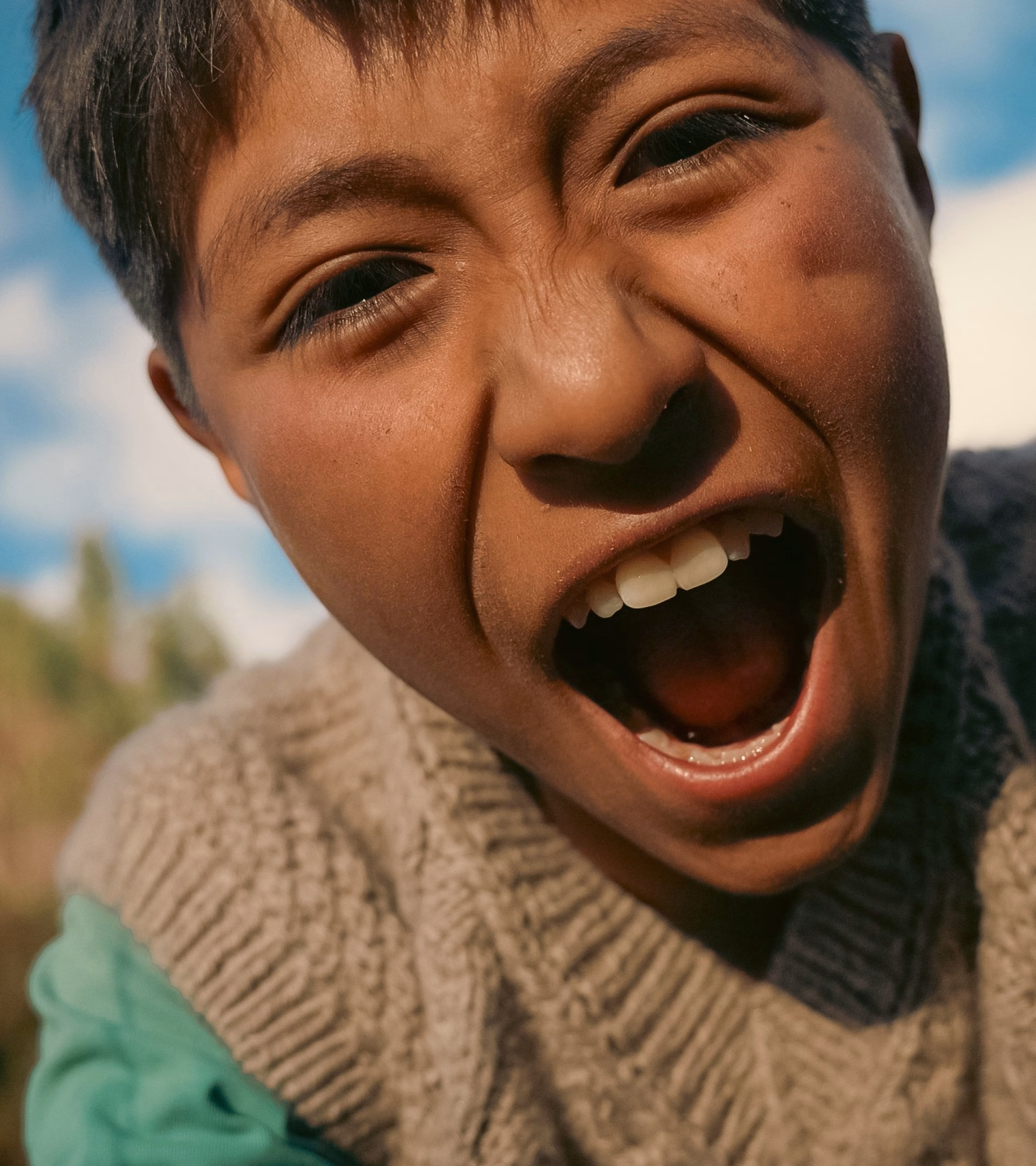 A close-up of a boy laughing joyfully, wearing a textured cream sweater against a bright sky backdrop.