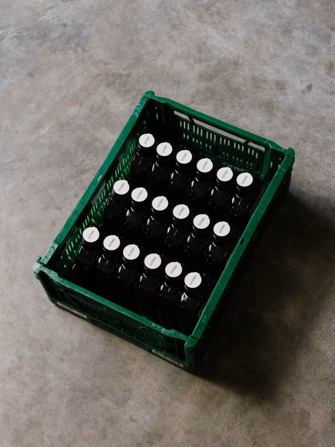 A green plastic crate holds black bottles with minimalist white labels, part of the Oraio design project.