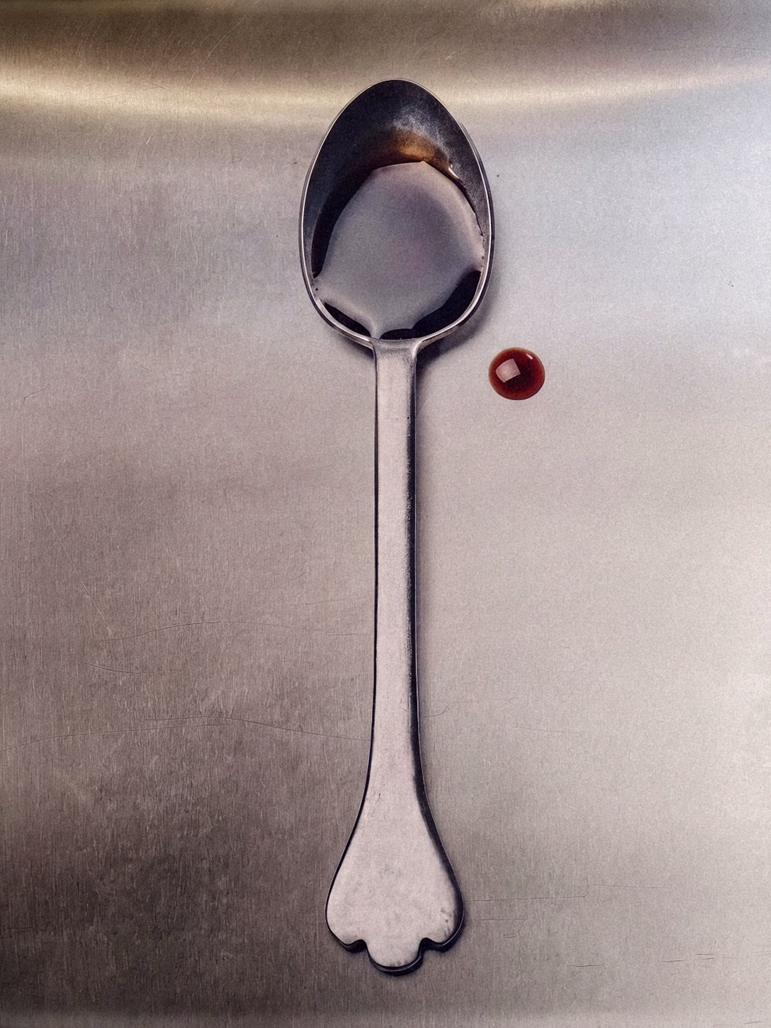 A tarnished silver spoon rests on a brushed metal surface, with a droplet of dark sauce beside it, showcasing minimalist design.