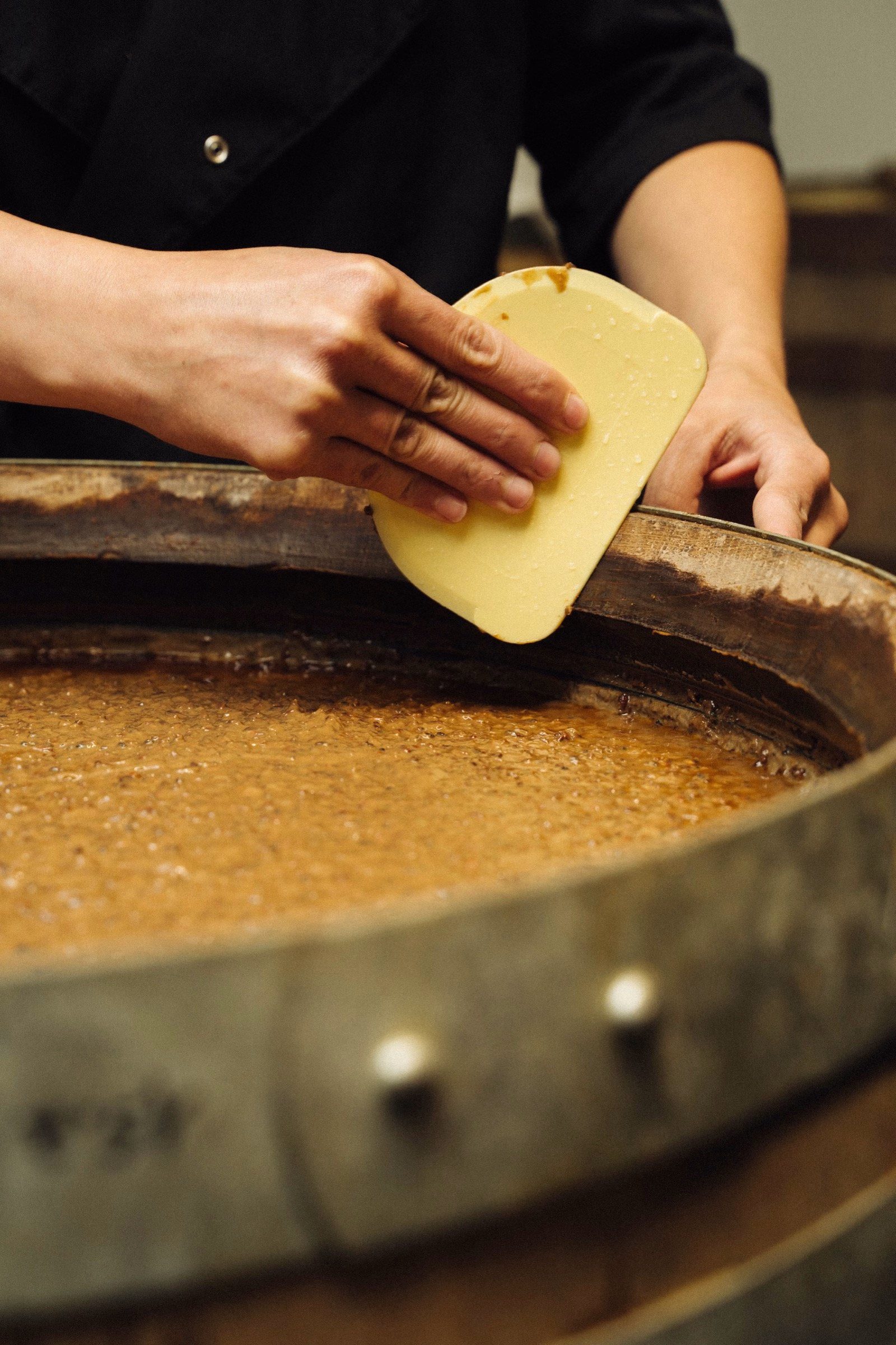 A hand spreads a yellow substance on the wooden edge of a barrel, showcasing Oraio's artisanal approach to food design.