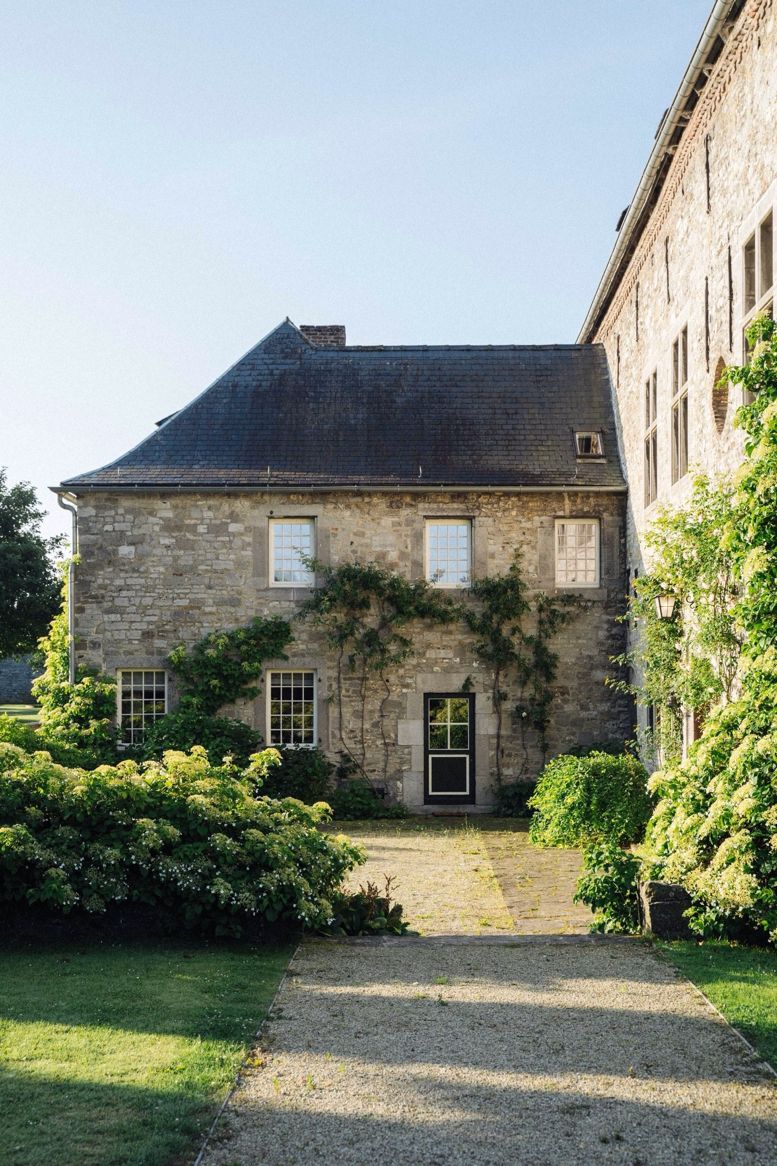 A rustic stone building with a slate roof is surrounded by lush green plants in an inviting outdoor space.