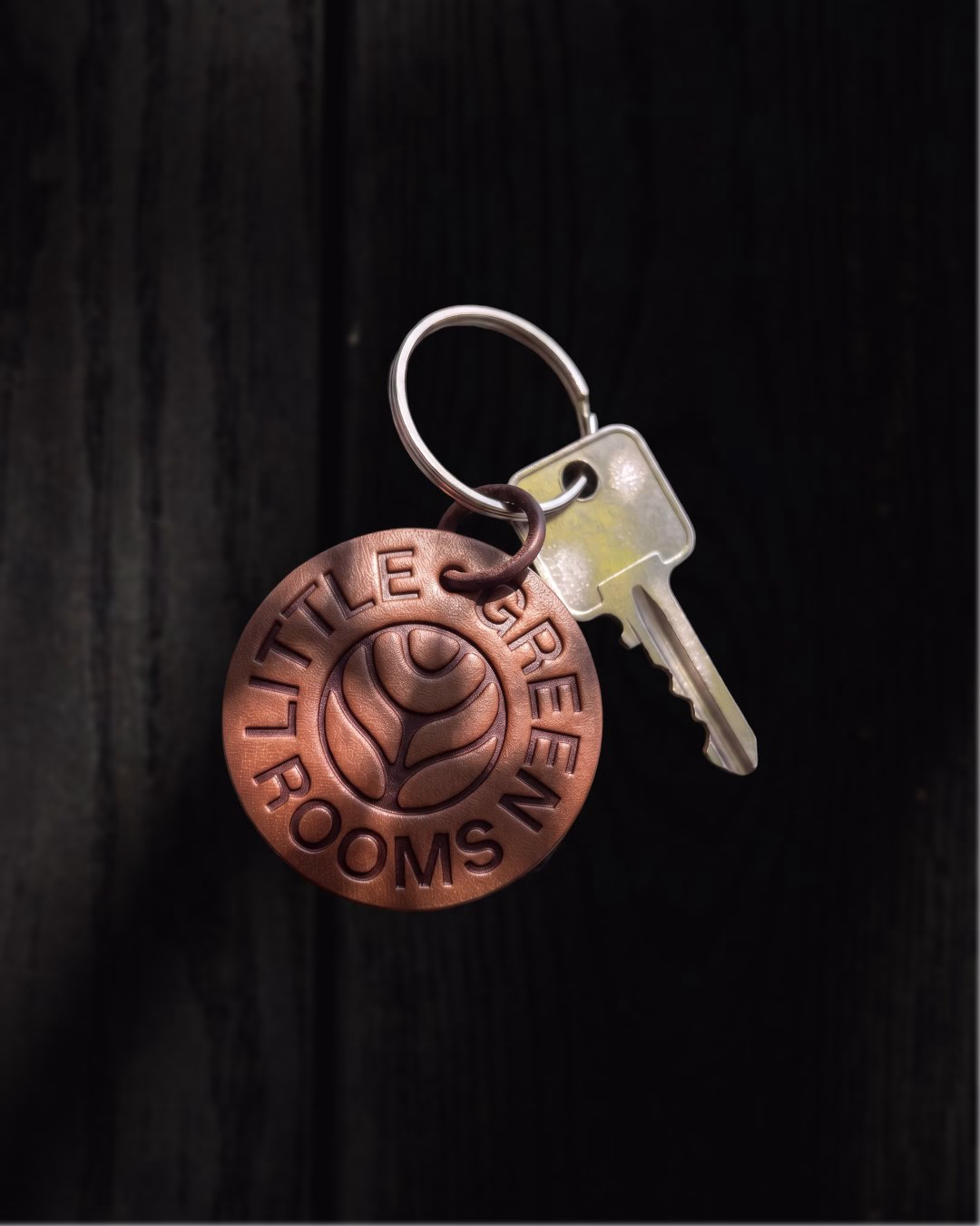 A copper keychain displaying "Little Green Rooms" alongside a silver key, set against a dark wooden background.