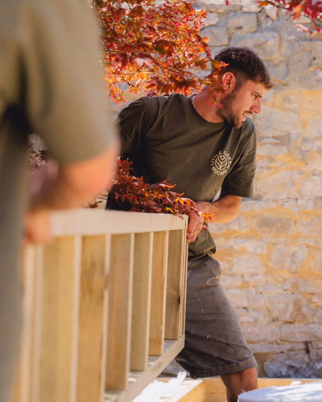 A man in a green shirt leans against a wooden railing, framed by vibrant red leaves in a stone-walled setting.