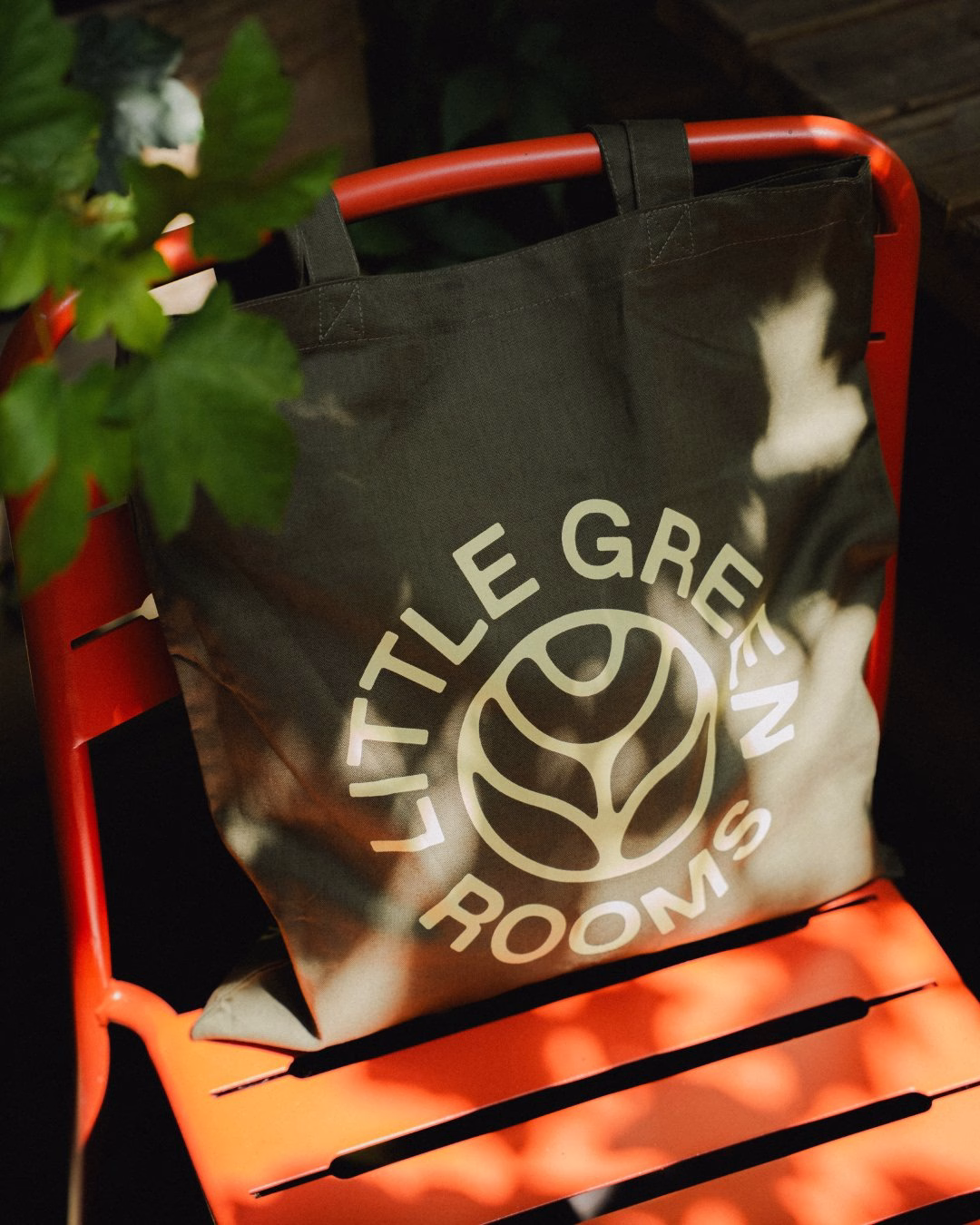 A green tote bag with the text "Little Green Rooms" rests on a vibrant orange chair, surrounded by leafy plants.