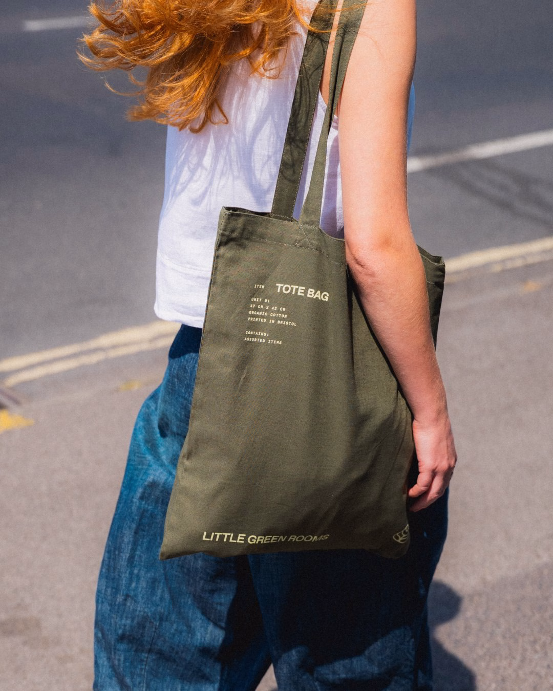 A person carries a khaki tote bag featuring the text "TOTE BAG" and "LITTLE GREEN ROOMS" in cream-colored typography.