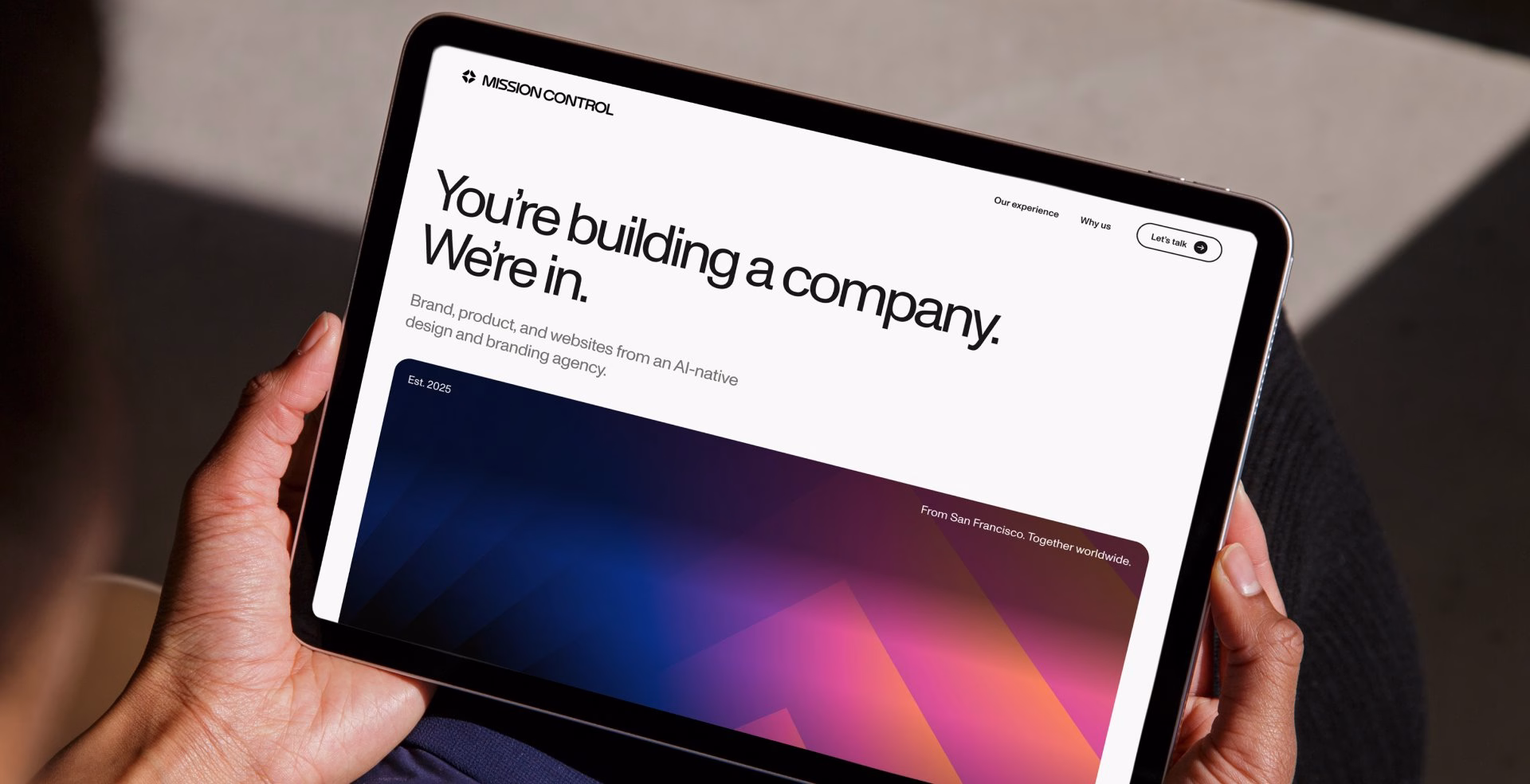 A hand holds a tablet displaying a modern website design with bold typography and a dusk gradient background.