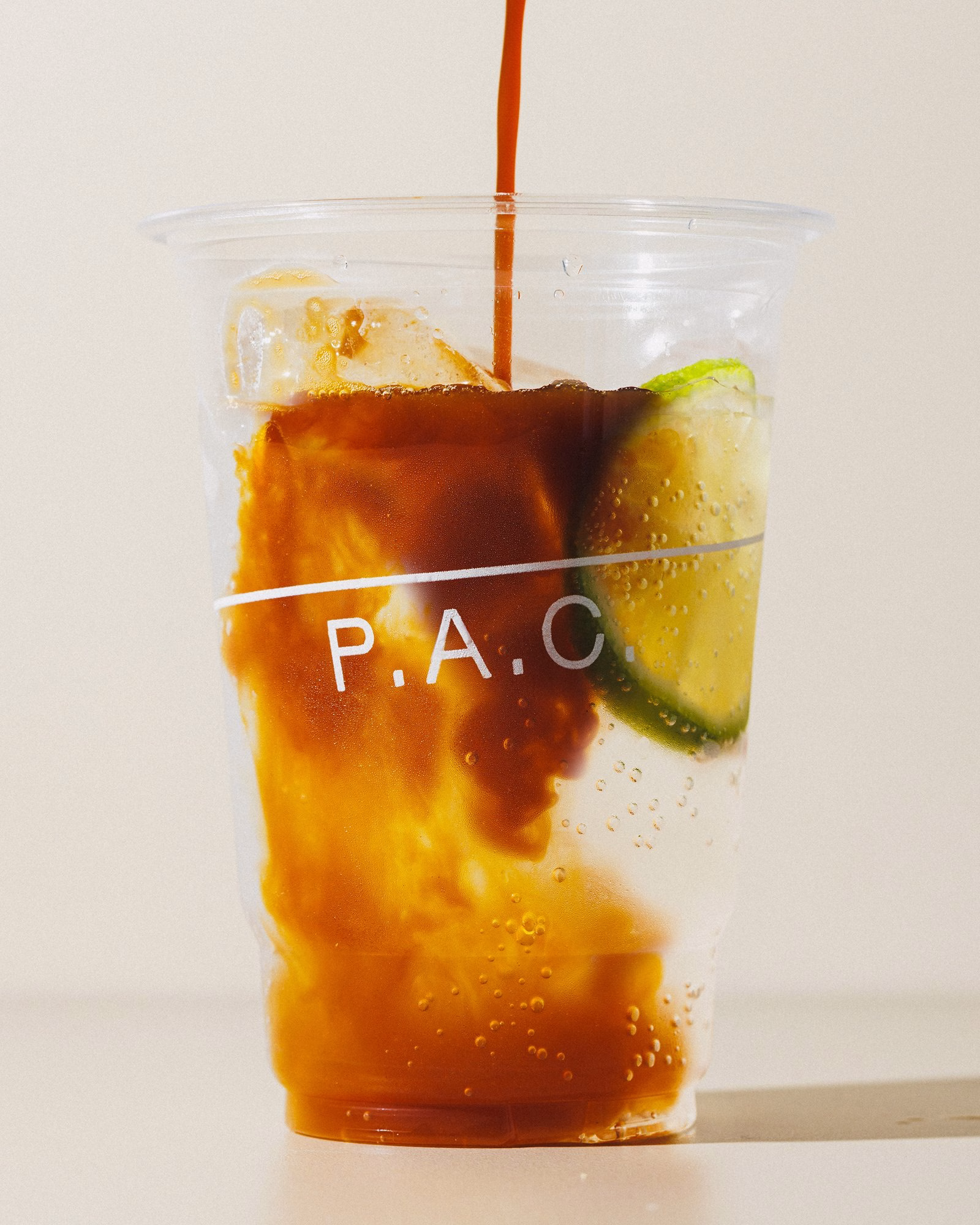A clear cup filled with a fizzy brown drink, ice, and a lime wedge, featuring the brand name P.A.C. in sleek typography.