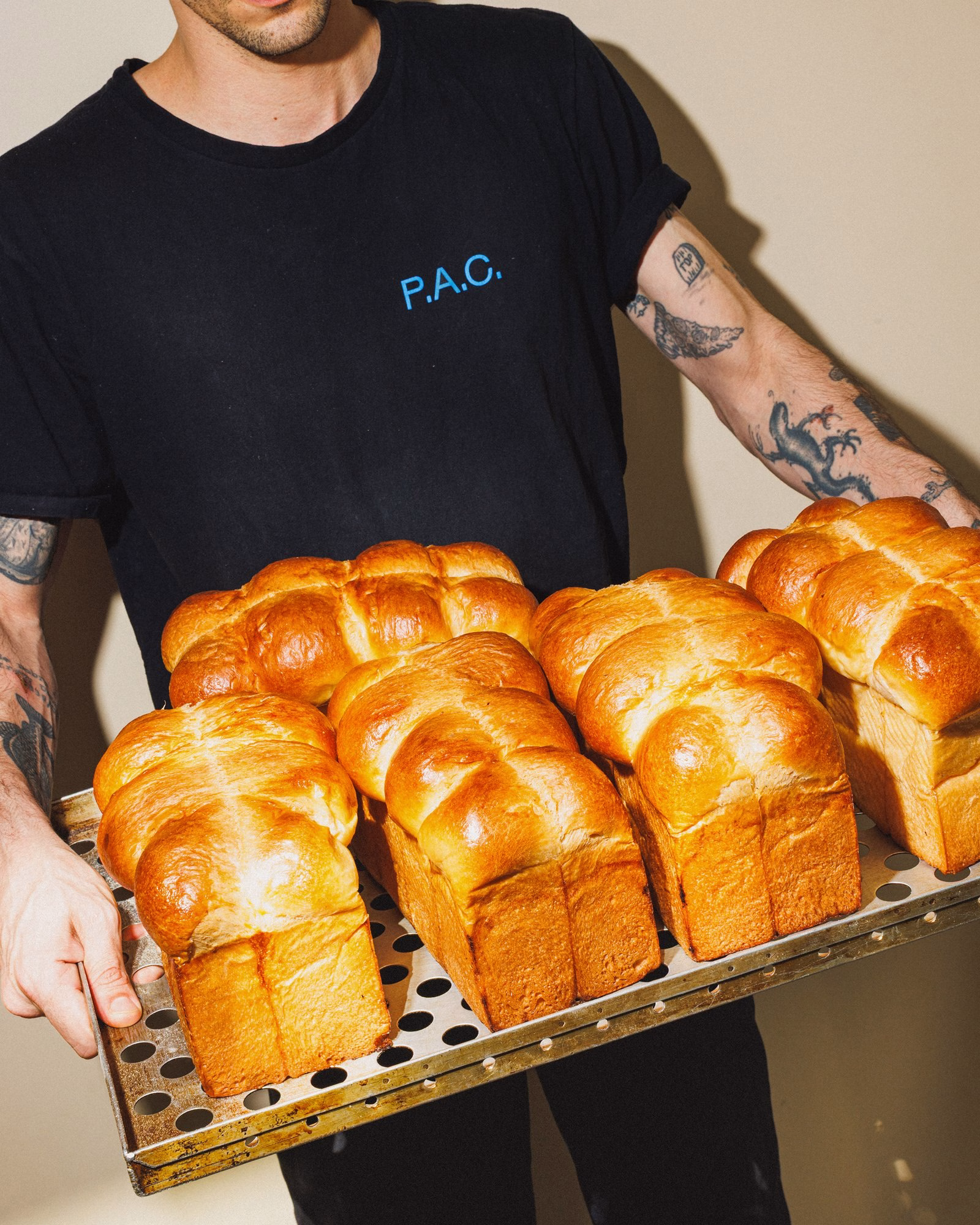 A person in a black shirt with "P.A.C." holds a tray of golden-brown loaves, showcasing bakery branding through color and form.