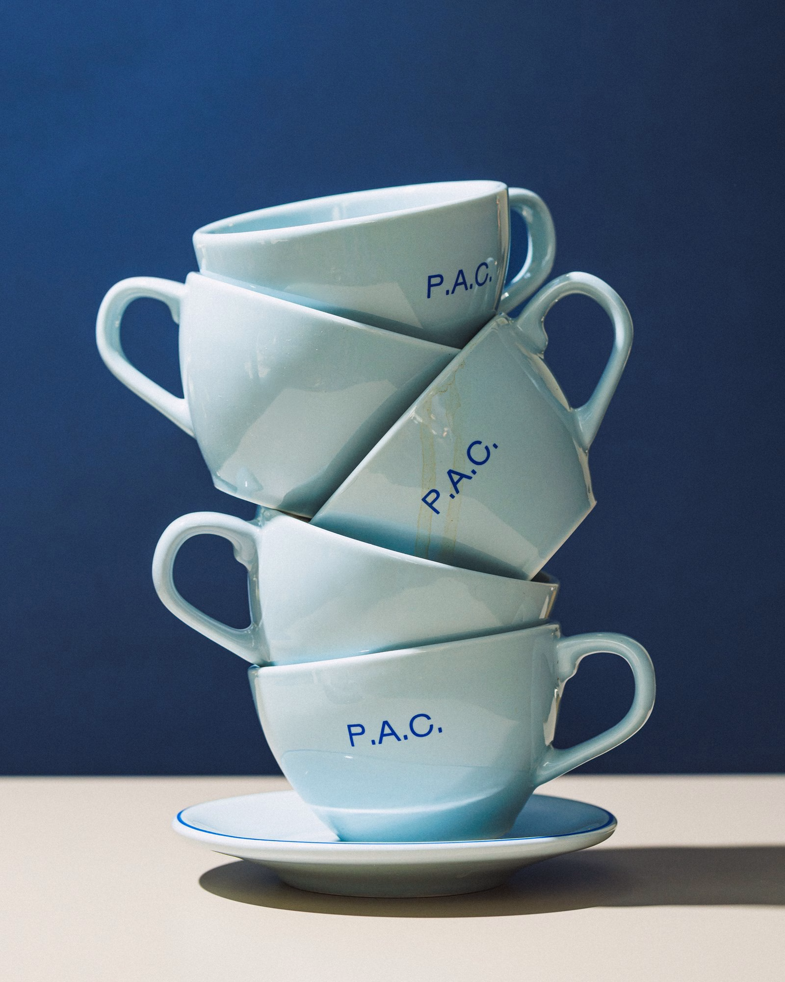 Stacked light blue coffee cups featuring "P.A.C." in bold blue typography against a dark blue background.