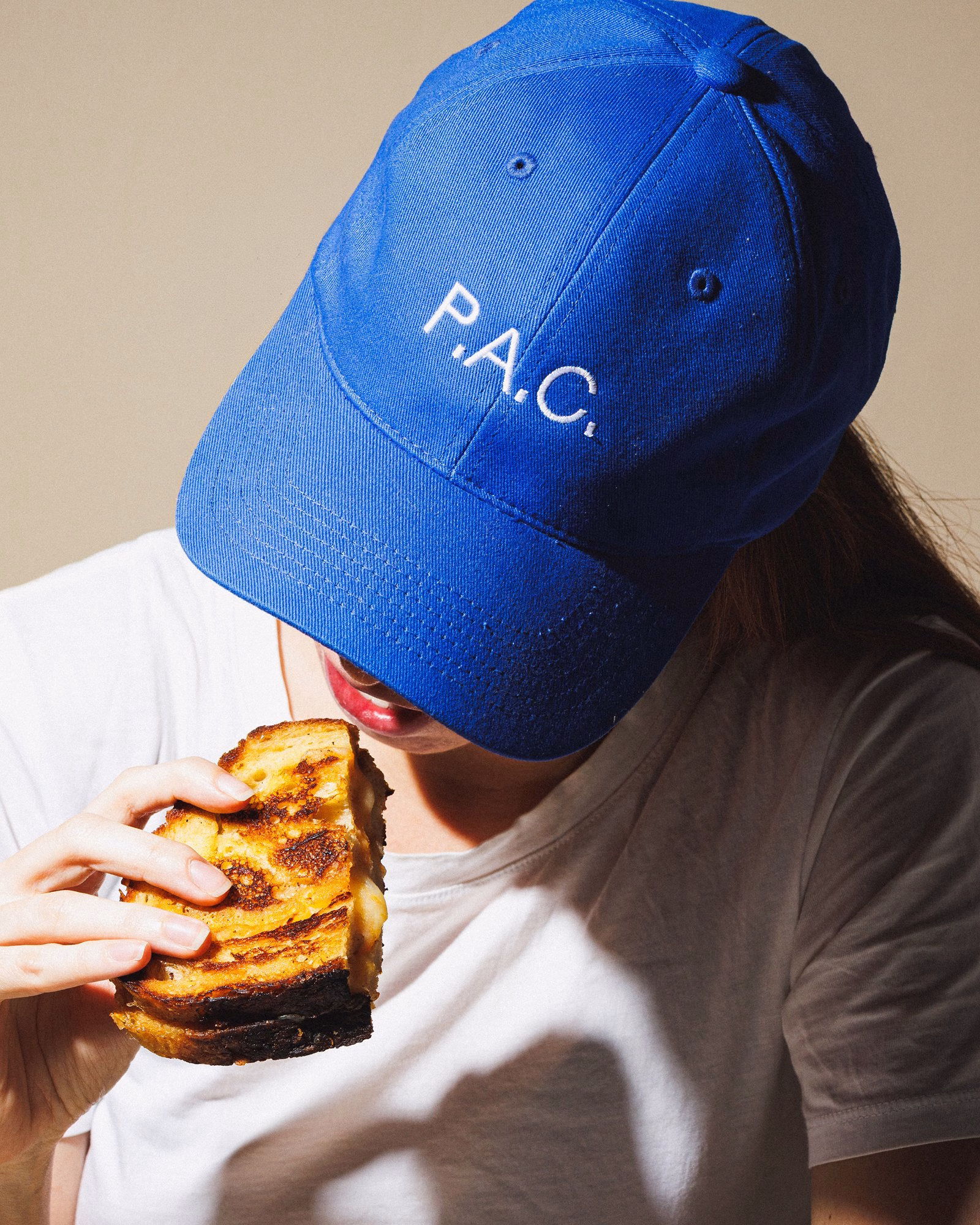 A person wears a blue cap with the brand name "P.A.C." while holding a grilled sandwich.