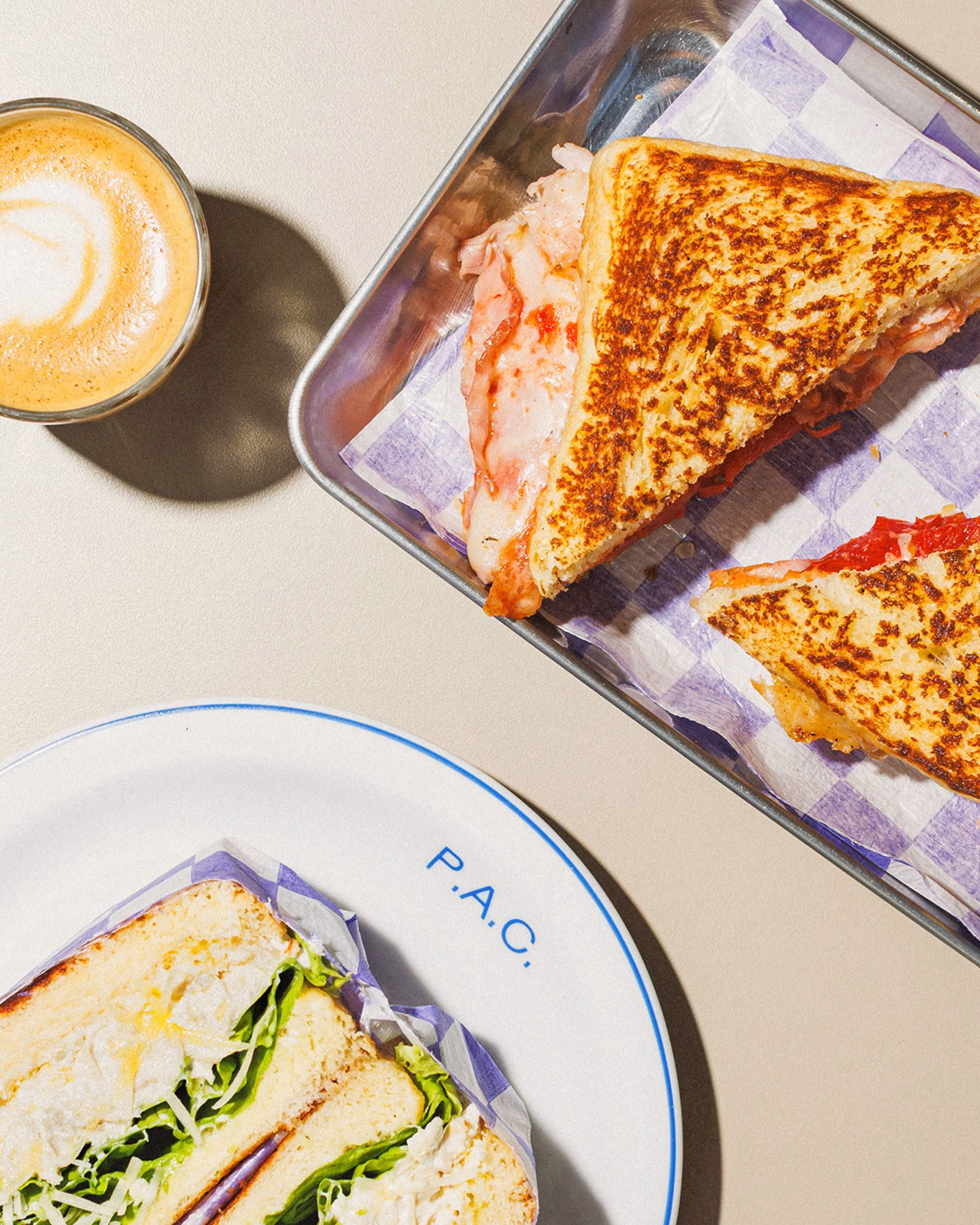 A plate with a sandwich featuring lettuce is displayed next to a tray of grilled sandwiches and a coffee cup.