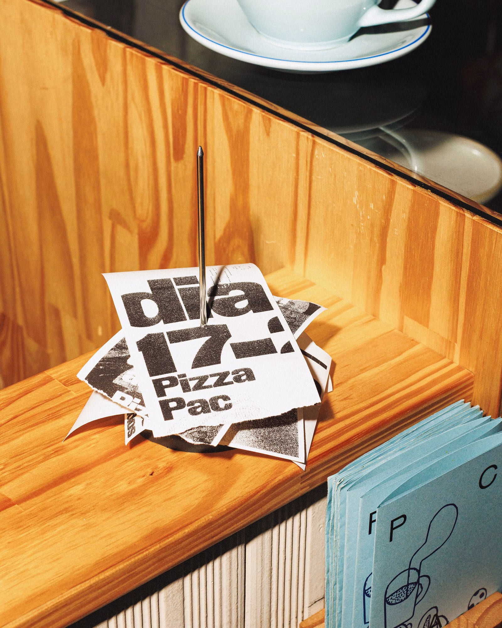 Stack of black and white printed cards labeled "Pizza Pac" on a wooden counter next to a light blue booklet and a coffee cup.