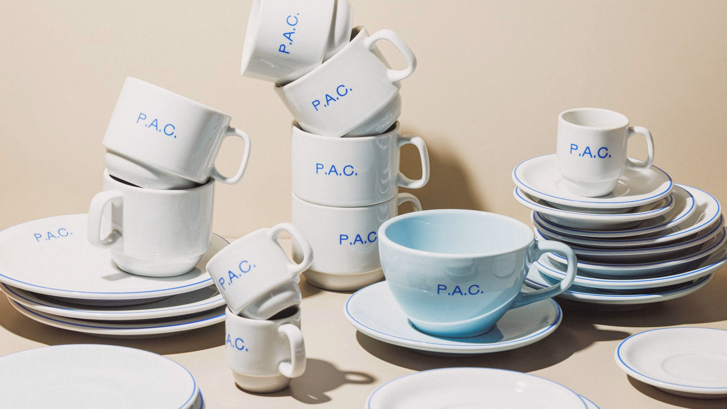 Stacked white and light blue ceramic cups and saucers feature the brand name "P.A.C." in blue typography against a neutral background.