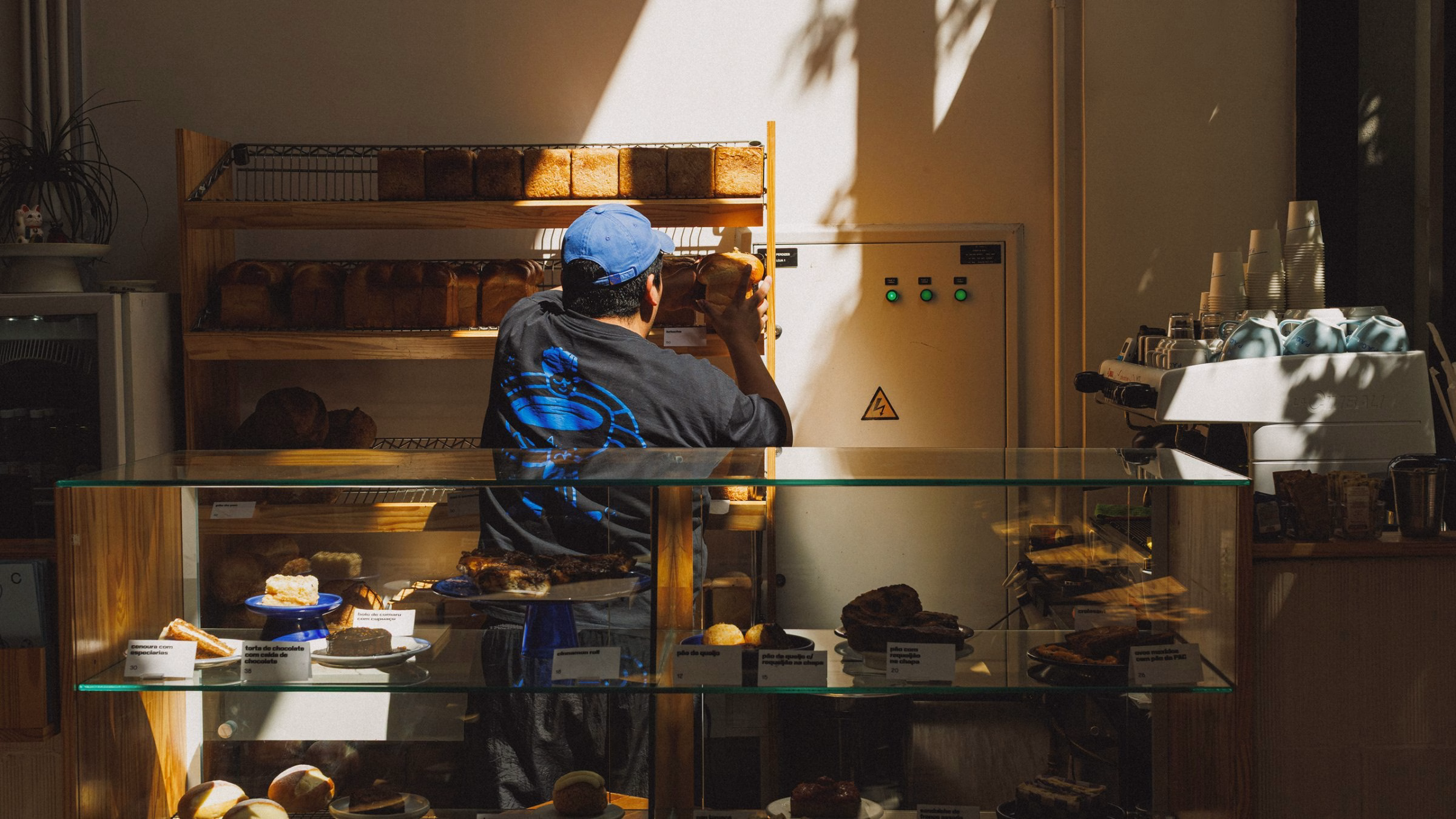 A baker wearing a black shirt with blue graphics arranges loaves on wooden shelves lit by warm sunlight.