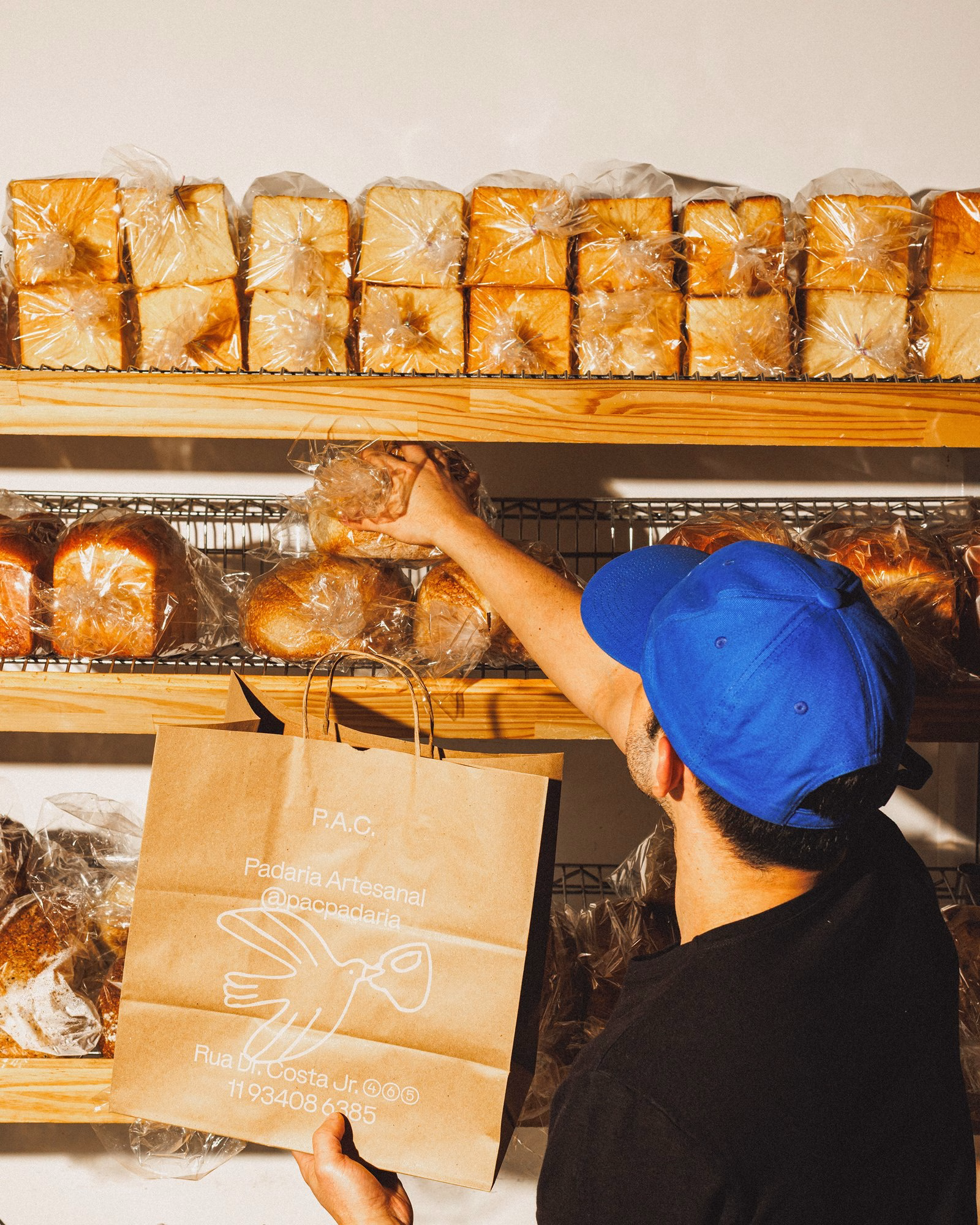 A person wearing a blue hat reaches for bread on a shelf, holding a brown paper bag with the P.A.C. logo and illustration.