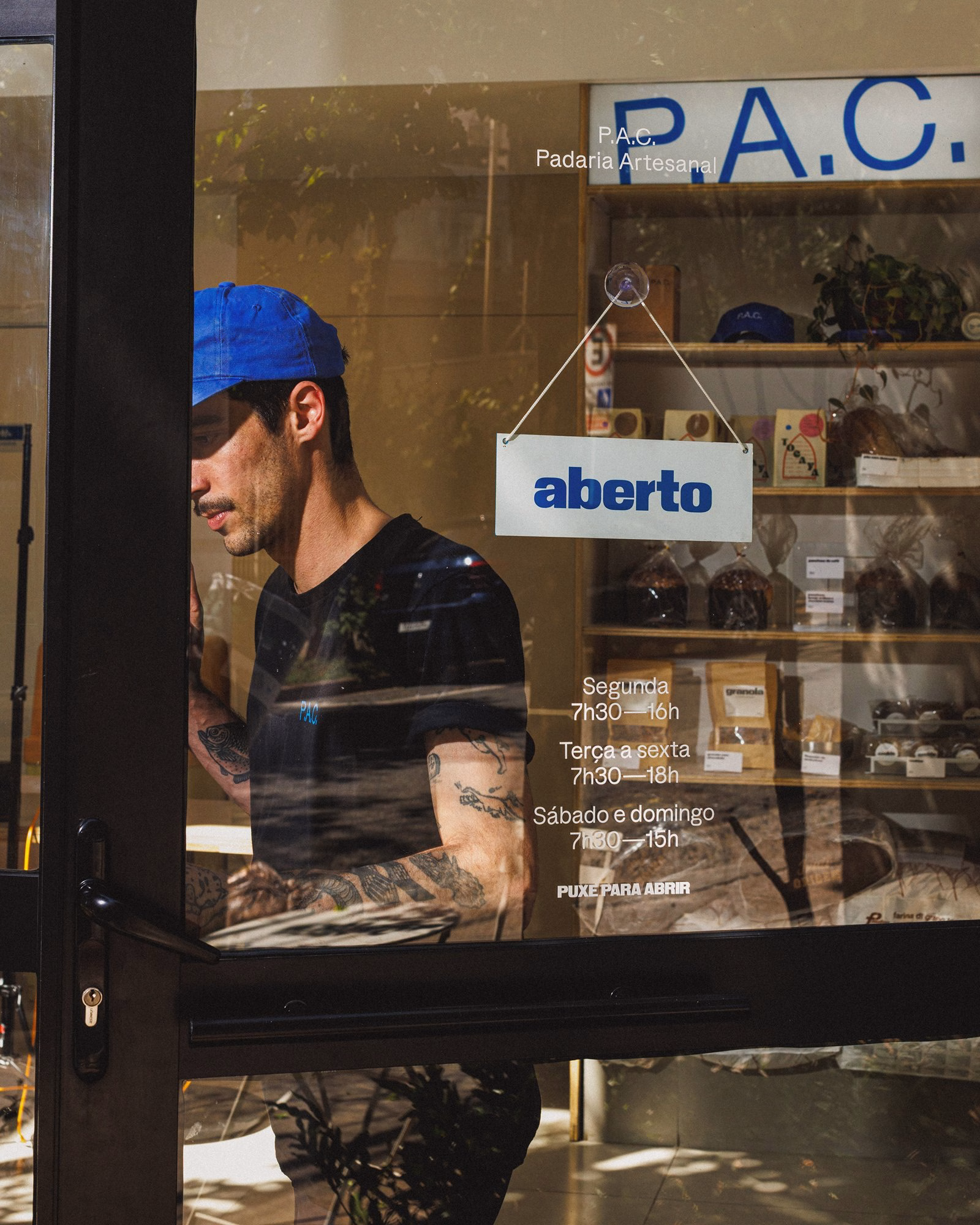 A bakery employee stands behind glass, showing a blue "aberto" sign and clean typography that enhances P.A.C.'s brand identity.