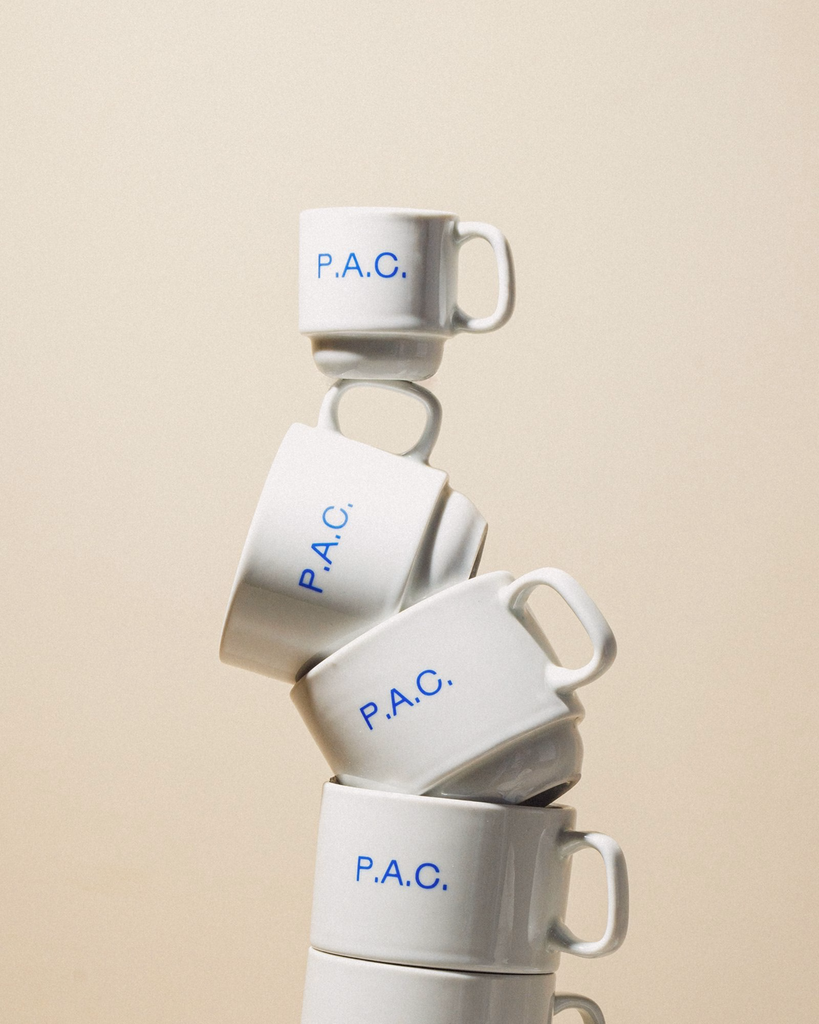Stacked white coffee mugs featuring the blue logo "P.A.C." against a soft beige background.