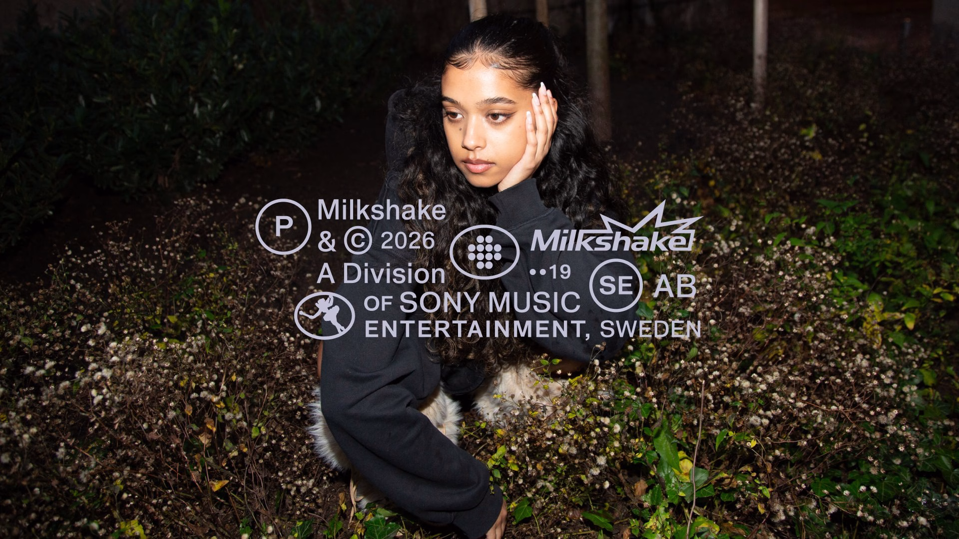 A young woman with curly hair poses thoughtfully among greenery, showcasing Milkshake's distinctive logo and branding.