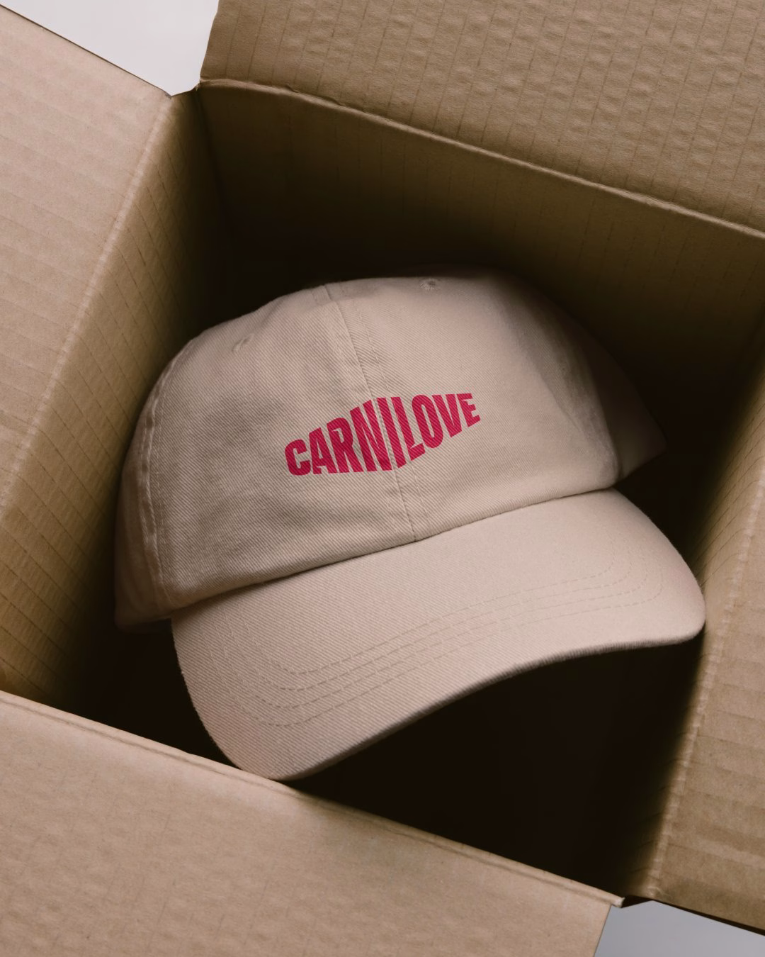A beige cap featuring bold red "CARNILOVE" text rests inside a cardboard box.