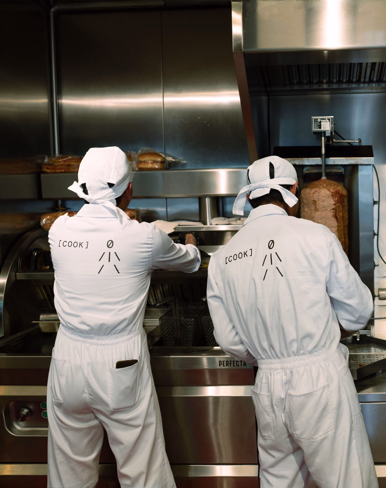 Two chefs in white uniforms with “[COOK]” printed on the back work at a stainless-steel döner cooking station.