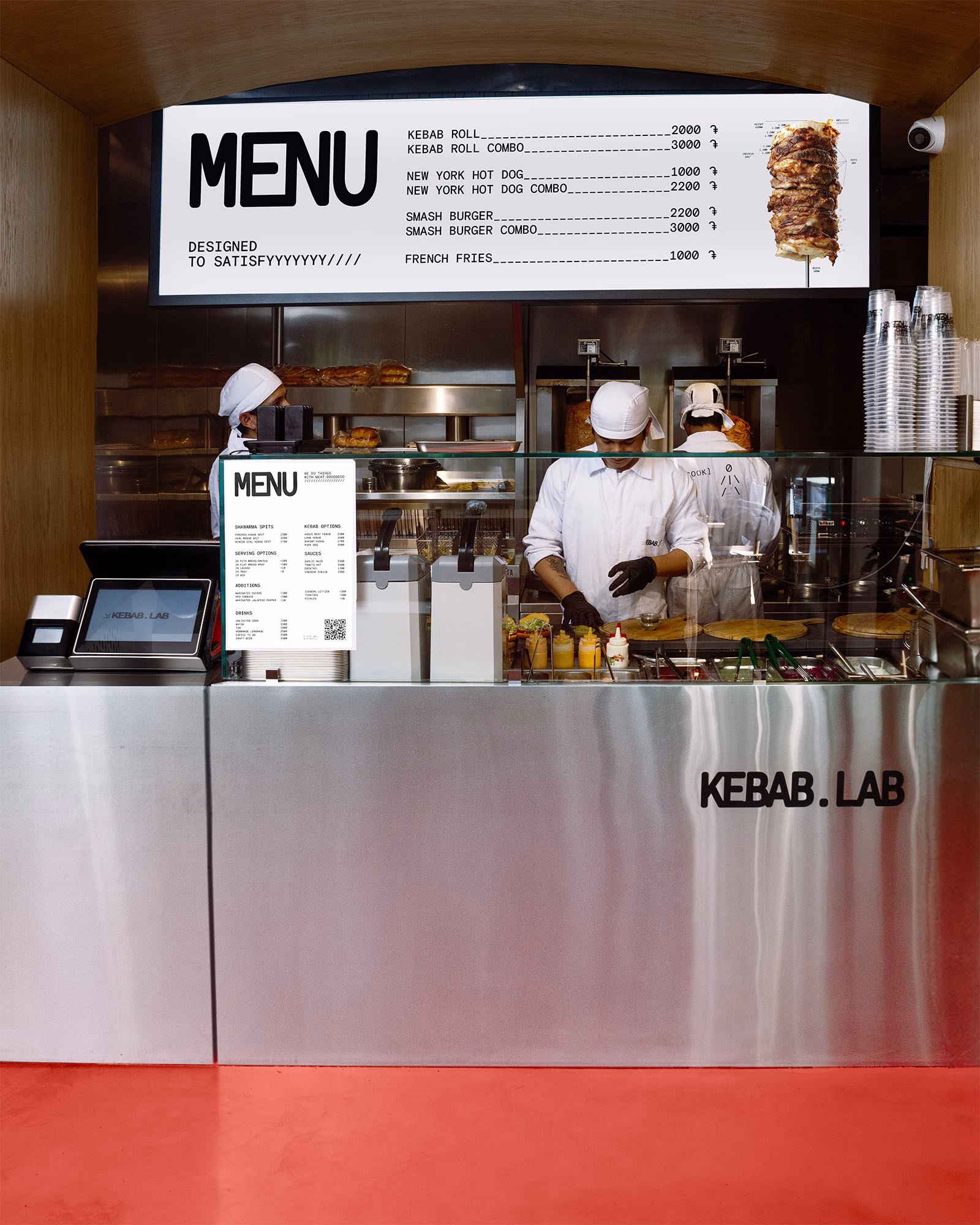 Stainless-steel service counter at Kebab.Lab featuring a menu board and chefs preparing food in white uniforms.
