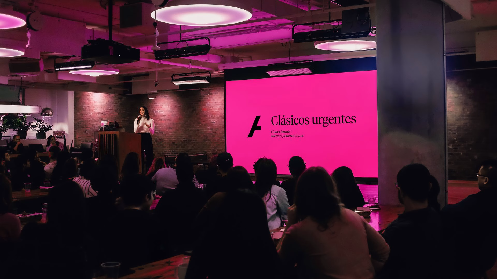 A speaker presents to an audience with a bright pink screen featuring the brand name "Clásicos urgentes" and modern typography.