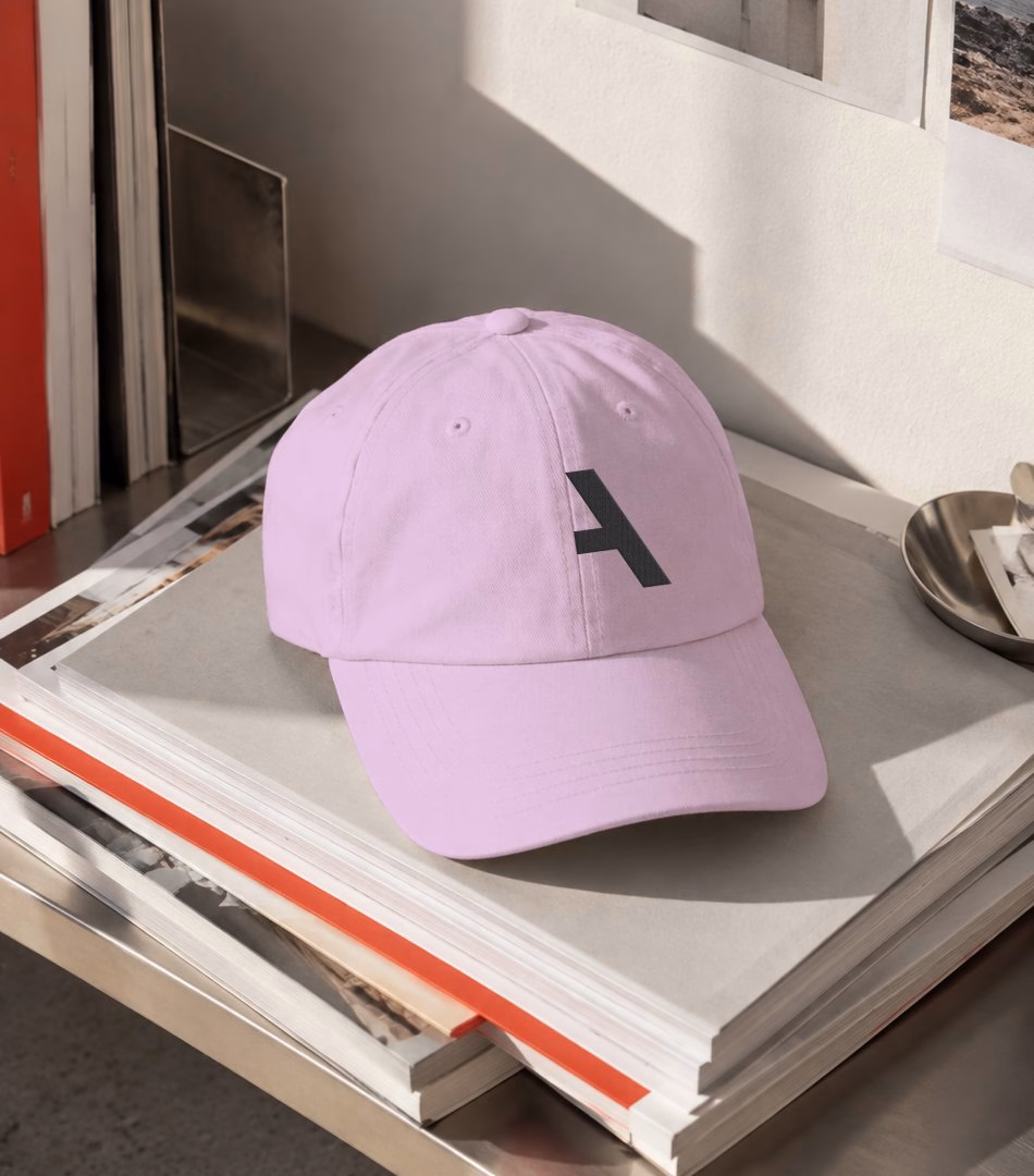 A pink cap with a stylized black "A" rests on a stack of books and magazines, illustrating the Alianza brand identity.