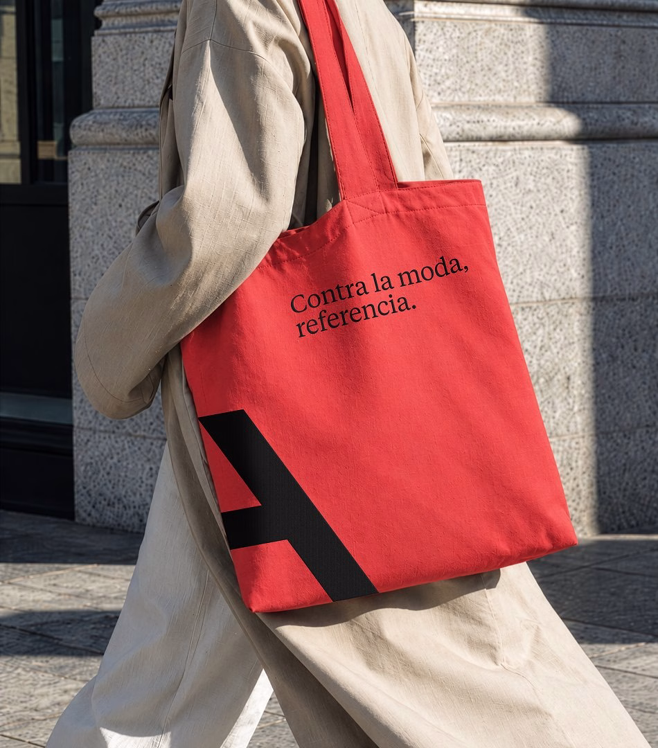 A person carries a red tote bag featuring the text "Contra la moda, referencia." and a large black letter A.