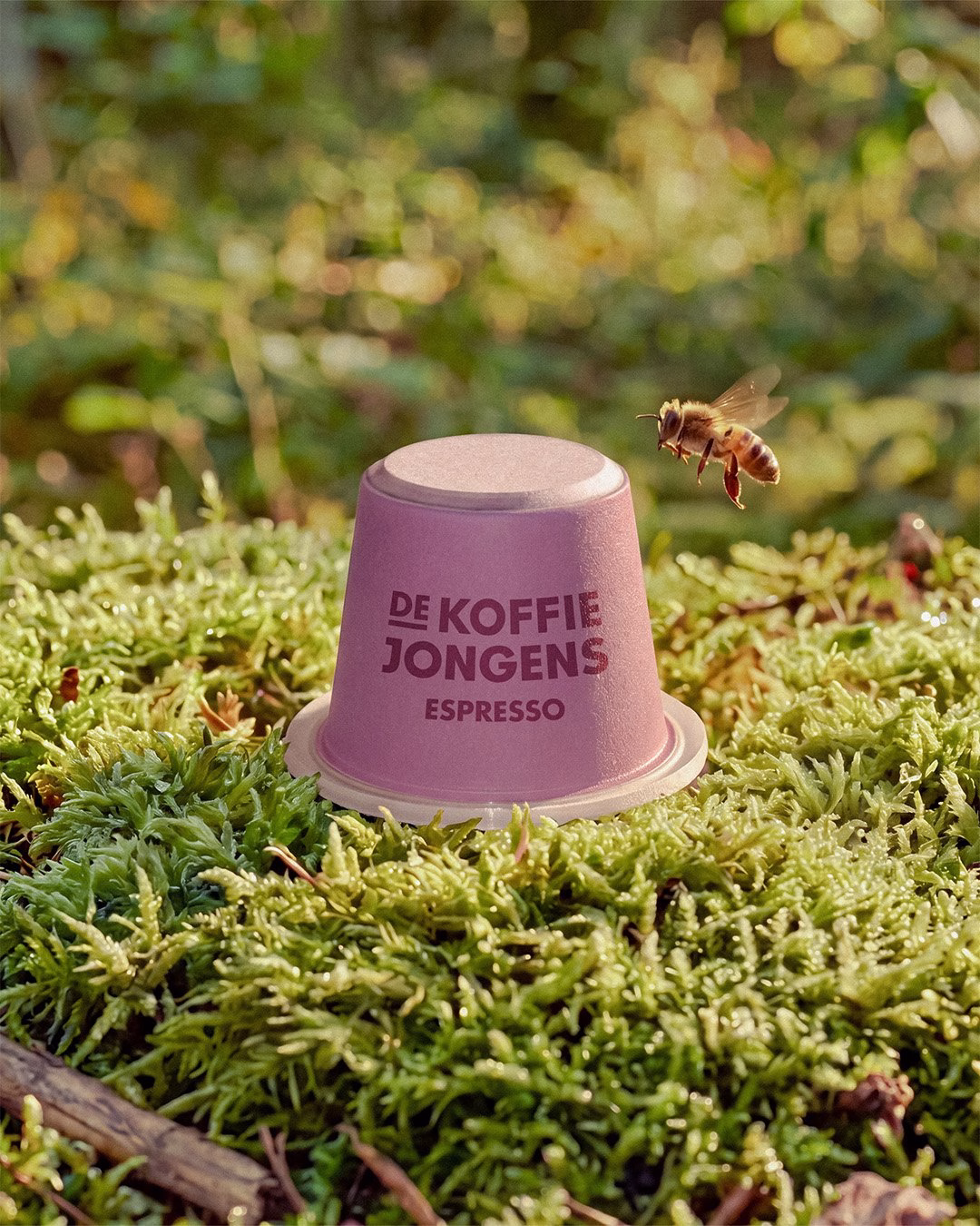 A pink coffee cup labeled "De Koffiejongens Espresso" sits atop green moss in a natural setting with soft light.
