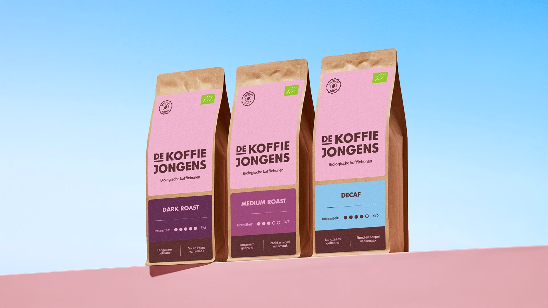 Three coffee bags from De Koffiejongens feature pink and brown packaging with distinct roast labels against a blue background.