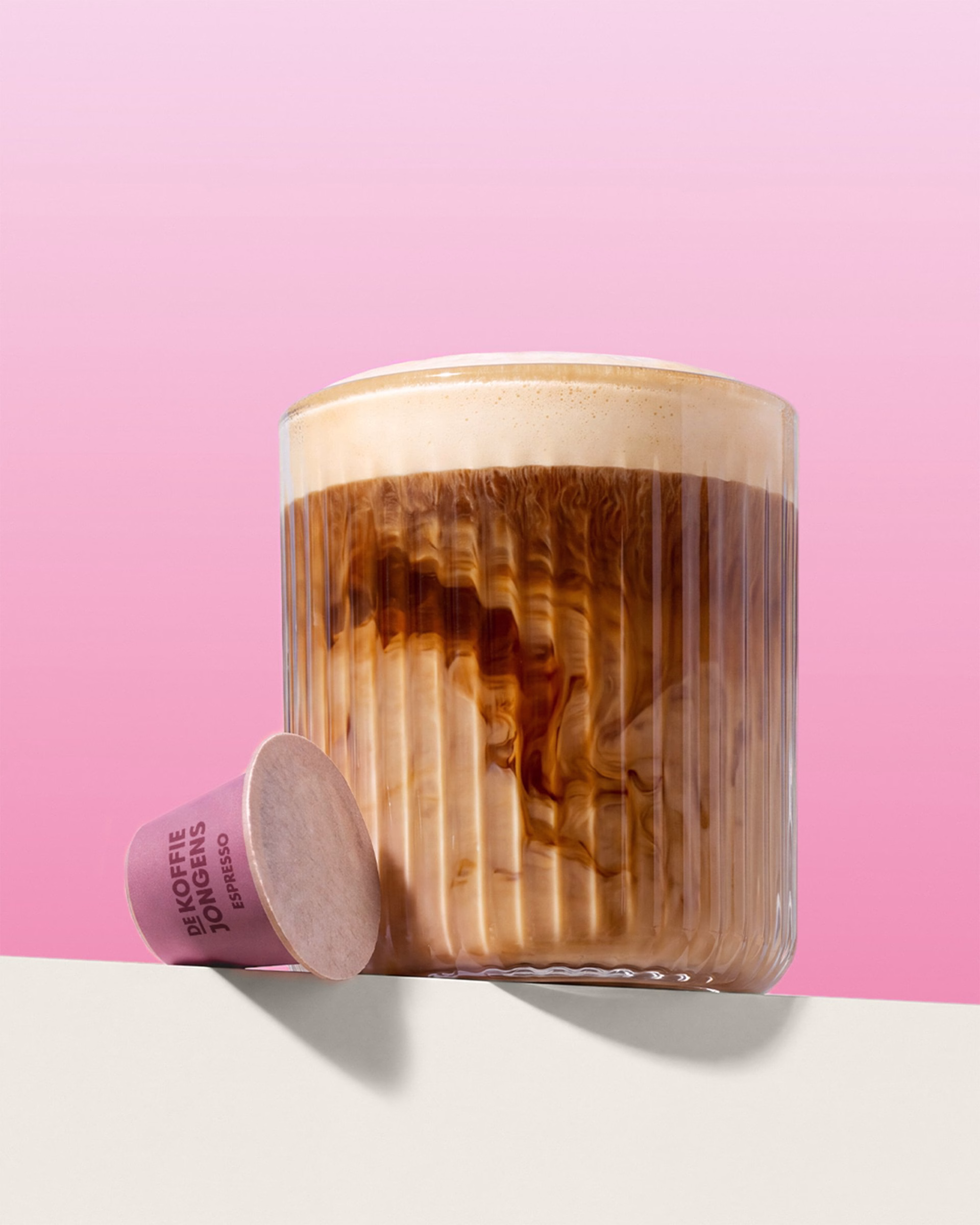 A clear glass with swirling coffee and foam sits next to a pink espresso capsule against a soft pink background.
