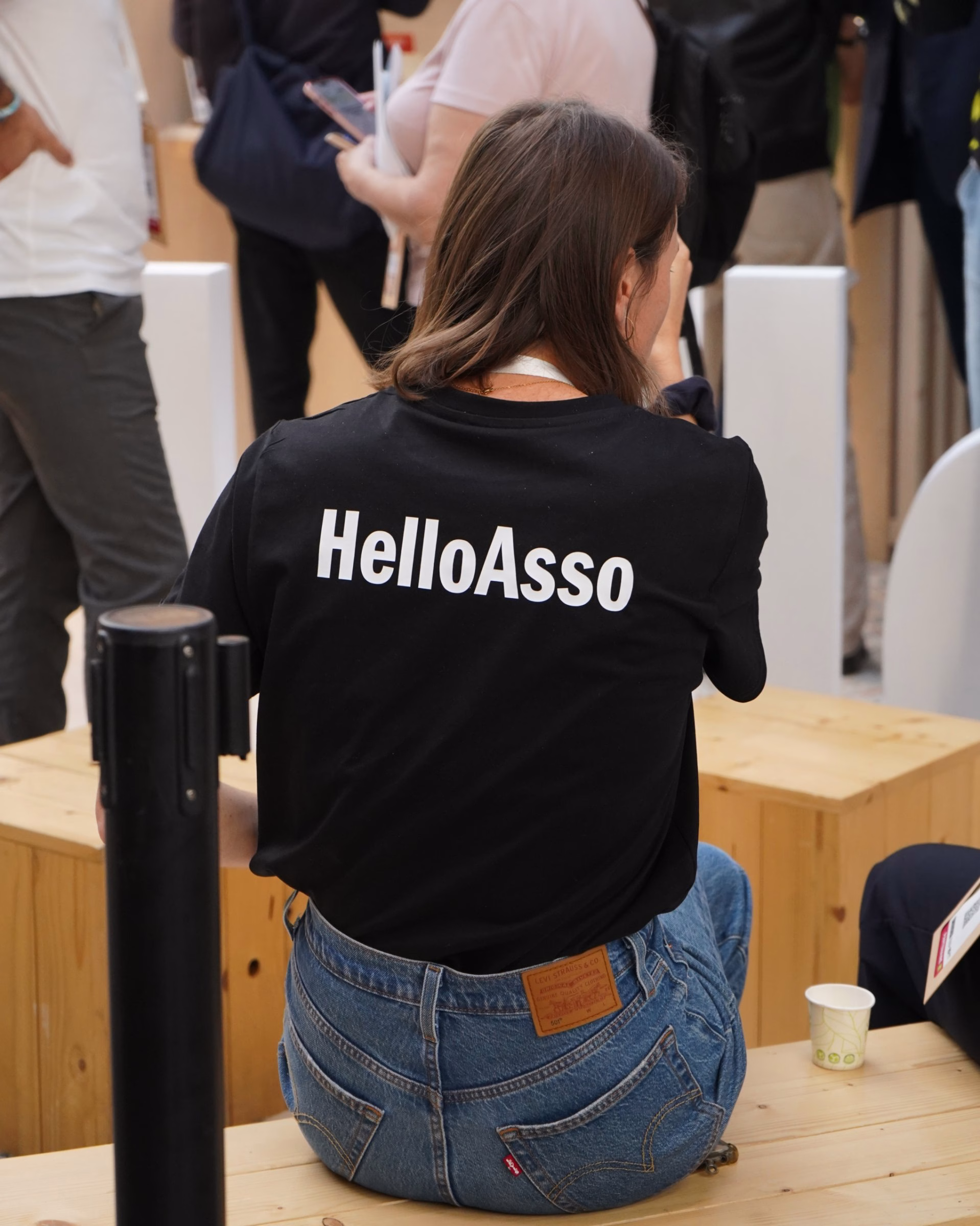A person seated on wooden benches wears a black shirt with "HelloAsso" printed in bold, white typography on the back.