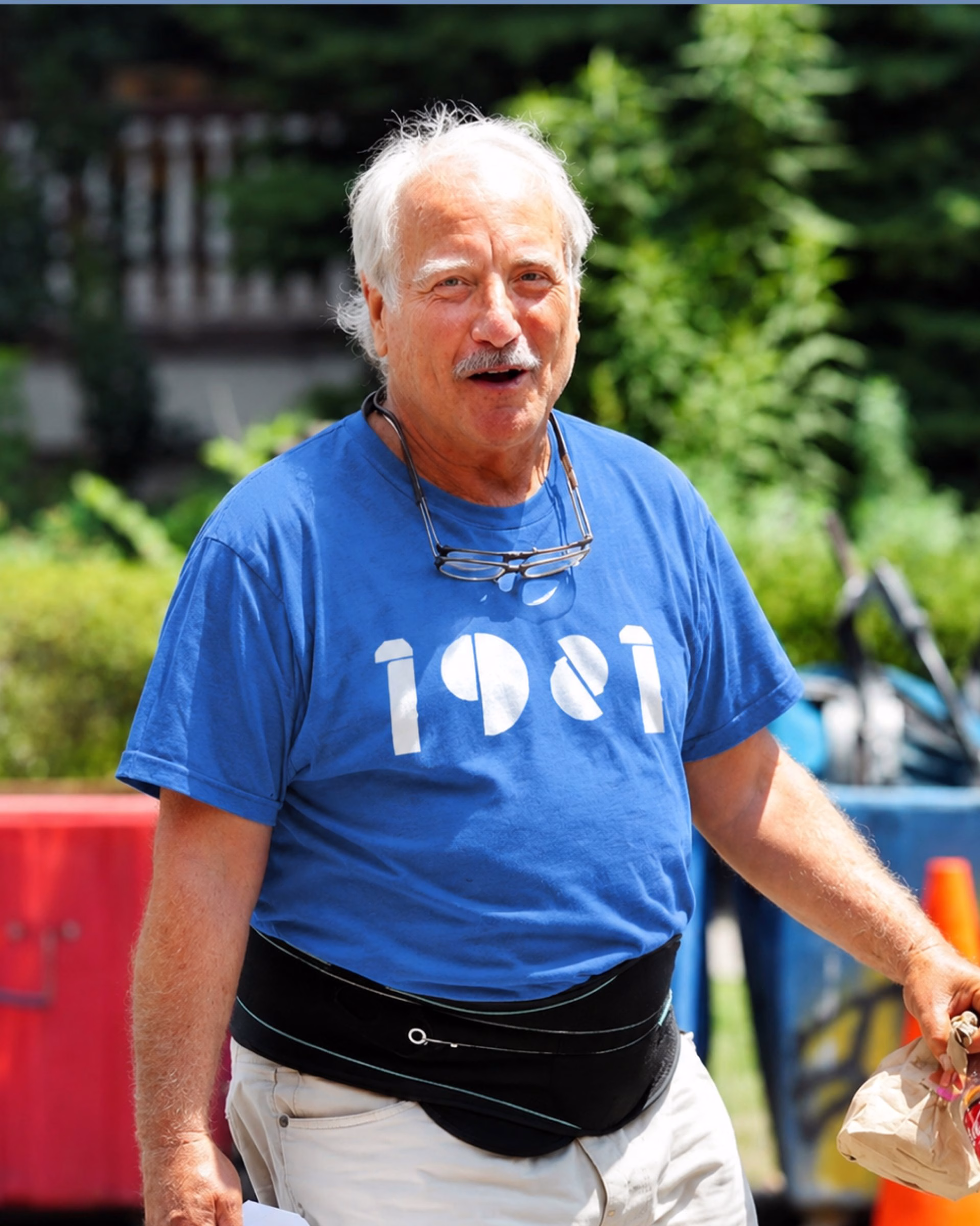 A man with white hair wears a blue shirt featuring bold white typography, smiling outdoors among greenery.