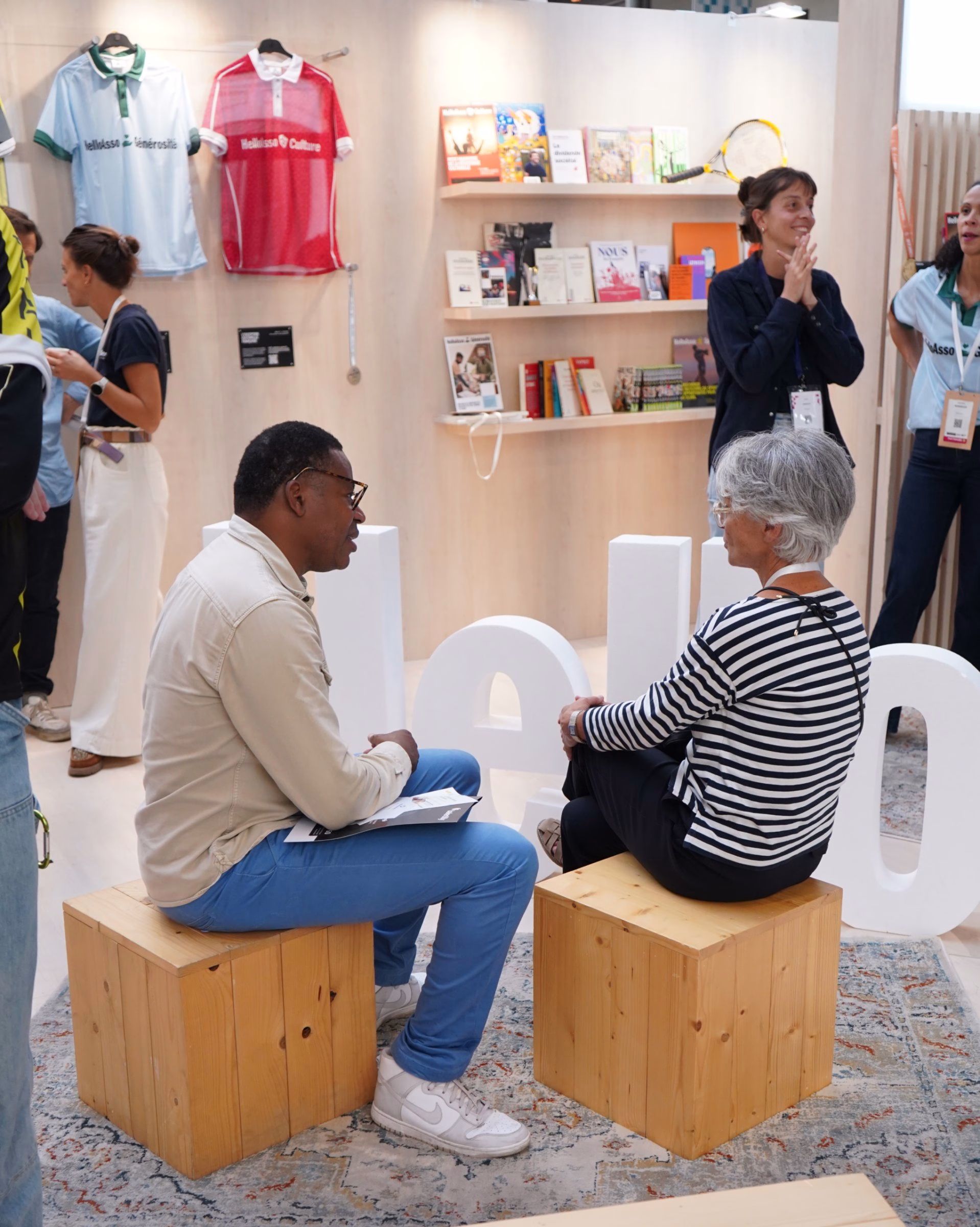 A man and a woman sit on wooden blocks, discussing in front of a light wooden display featuring books and jerseys.
