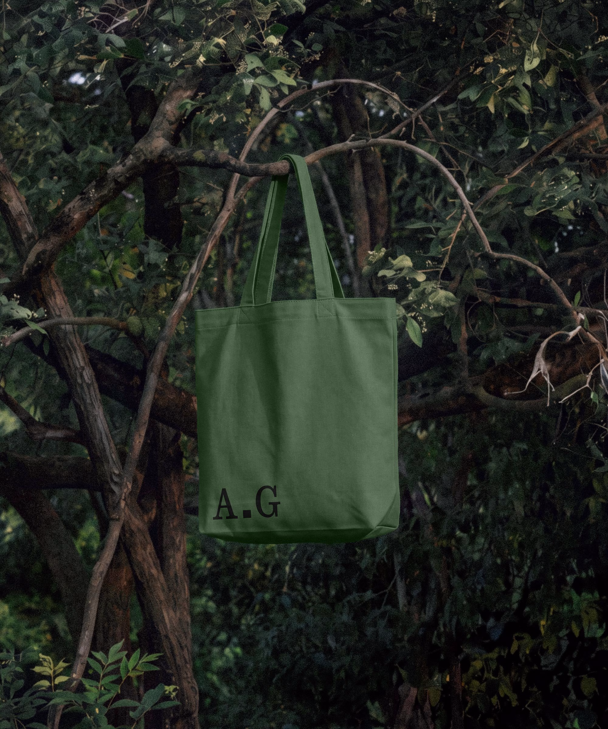A lichen green tote bag with the initials 'A.G' hangs from a branch in a lush, green forest setting.