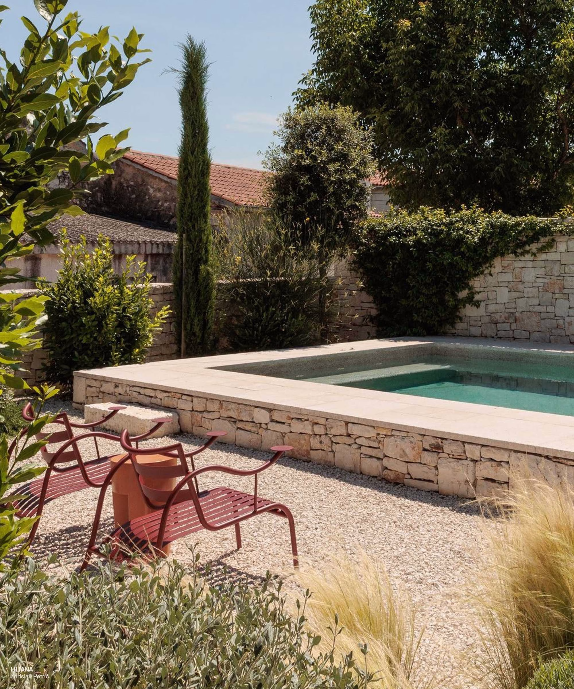A lichen green garden features a stone pool and red metal chairs amidst lush foliage and decorative grasses.