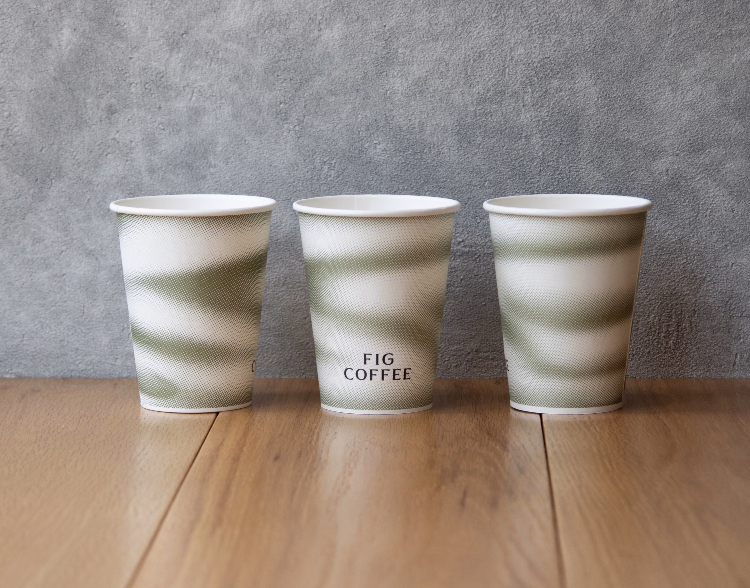 Three coffee cups feature a green gradient design with the text "Fig Coffee" near the base, set against a grey backdrop.