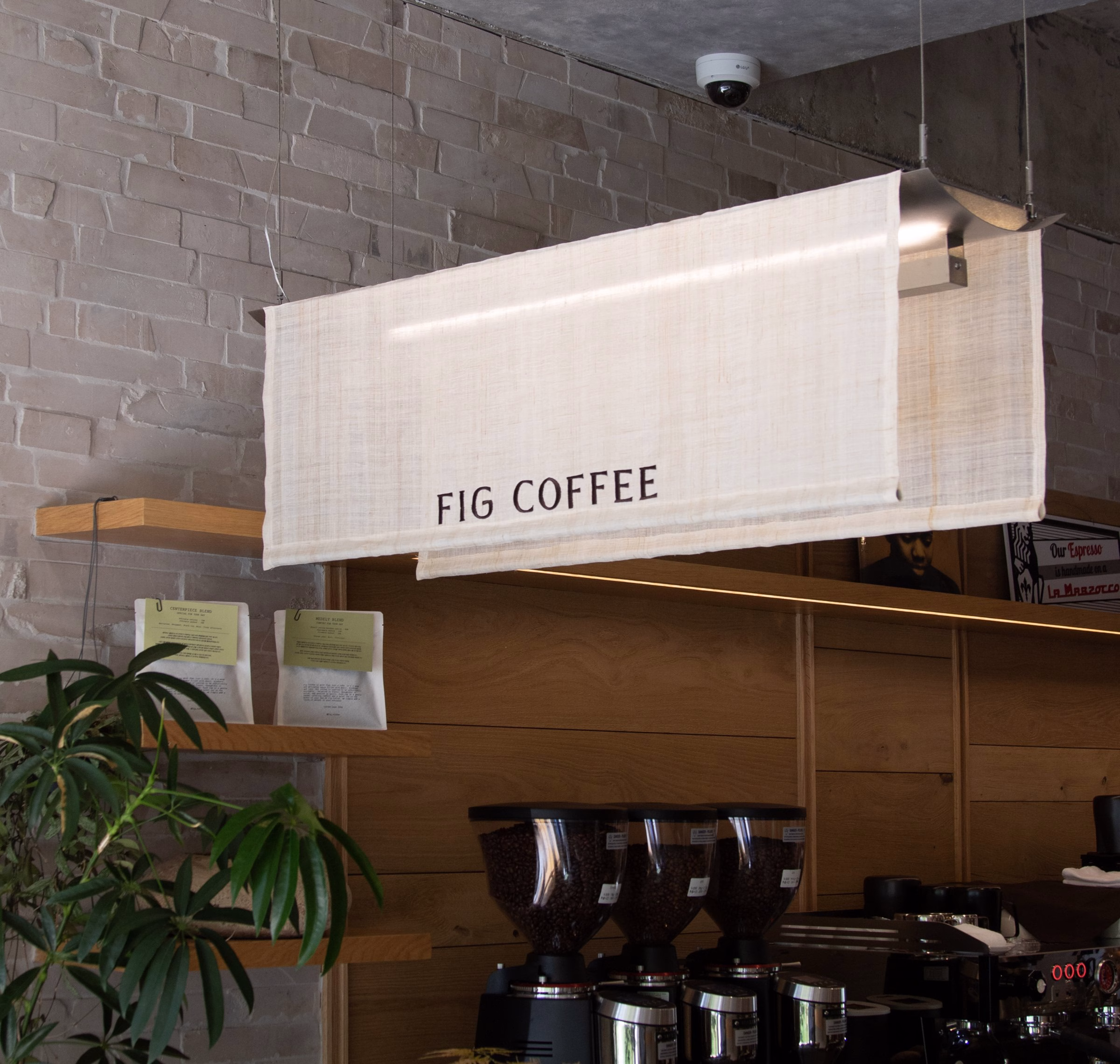 A light fabric sign reading "FIG COFFEE" hangs above wooden shelves and coffee grinders in a minimalist cafe setting.