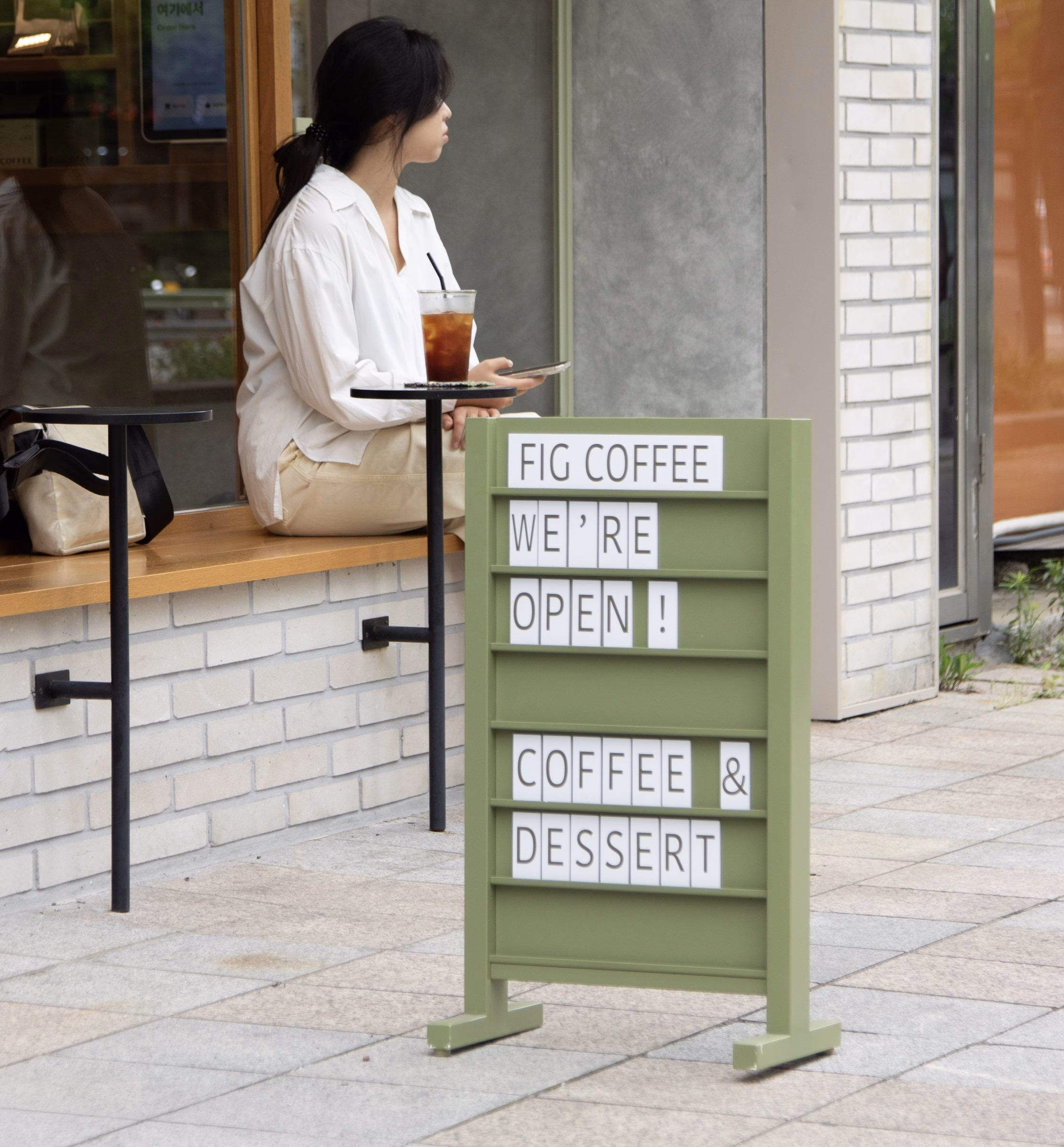 A woman sits by the window of Fig Coffee, next to a green sign advertising "Open" and "Coffee & Dessert."