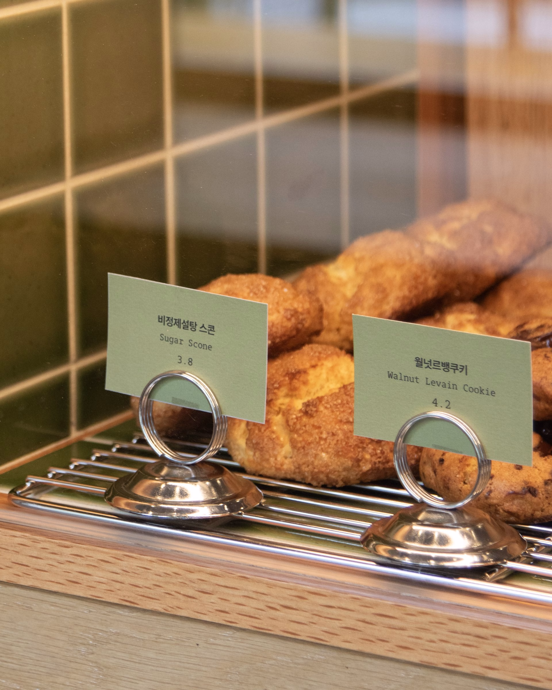 Green price tags display items in a cafe pastry case, highlighting a walnut levain cookie and sugar scone.