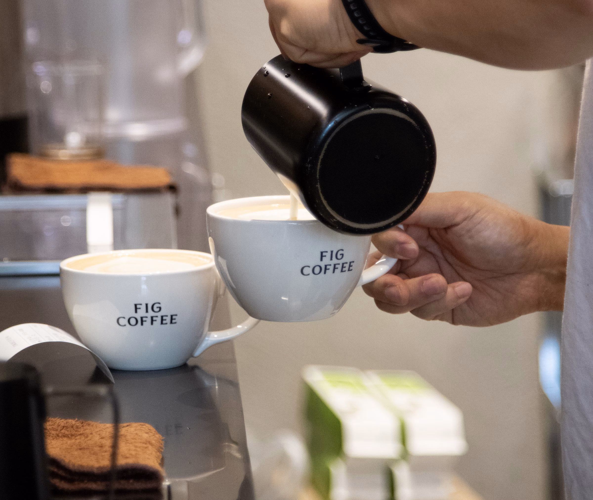 A hand pours milk into a white cup with "FIG COFFEE" printed on it, with another cup nearby and green packaging in the background.