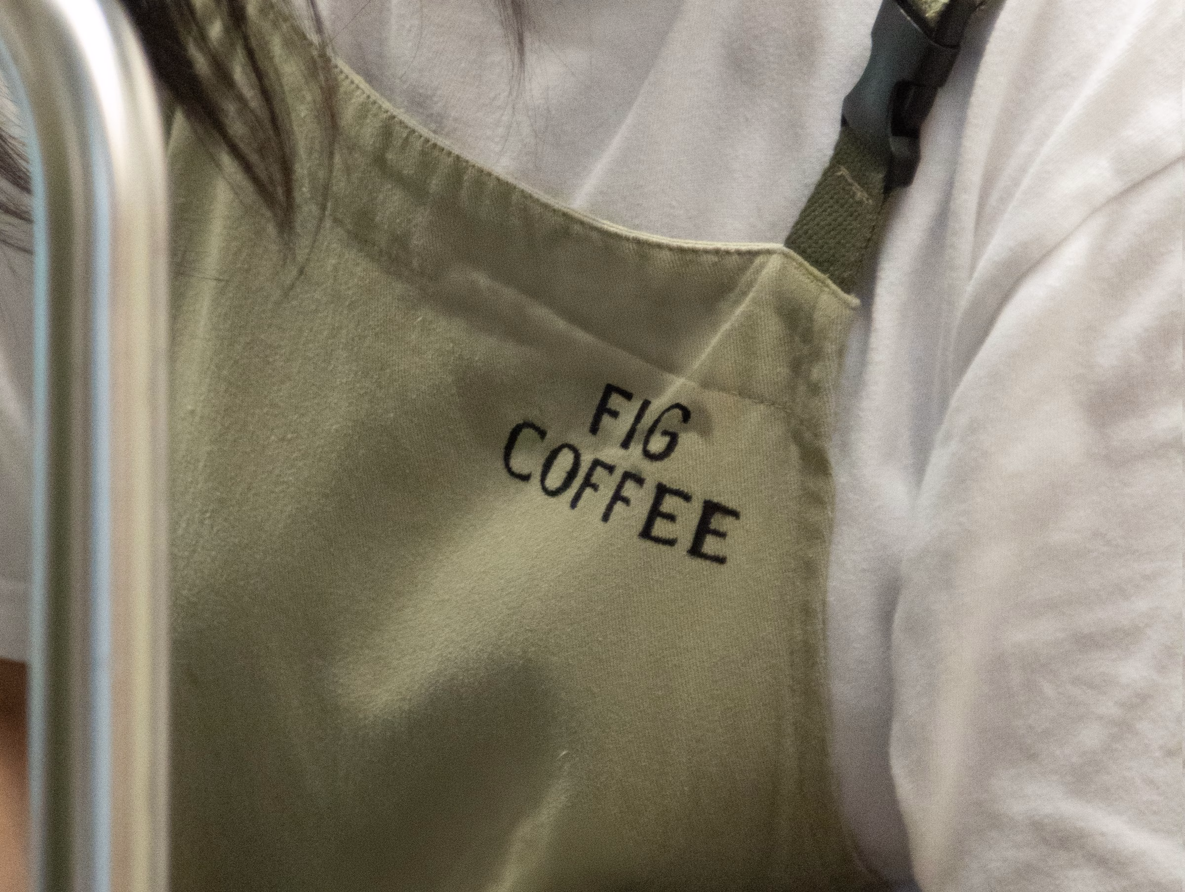 An olive green apron with "FIG COFFEE" embroidered in black, worn by a barista beside a white shirt.