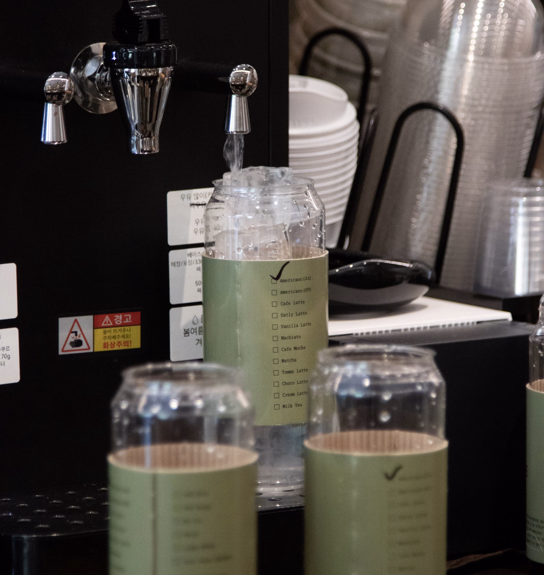 A coffee machine dispenses water into a clear jar with a green label listing drink options, showcasing minimalist design.