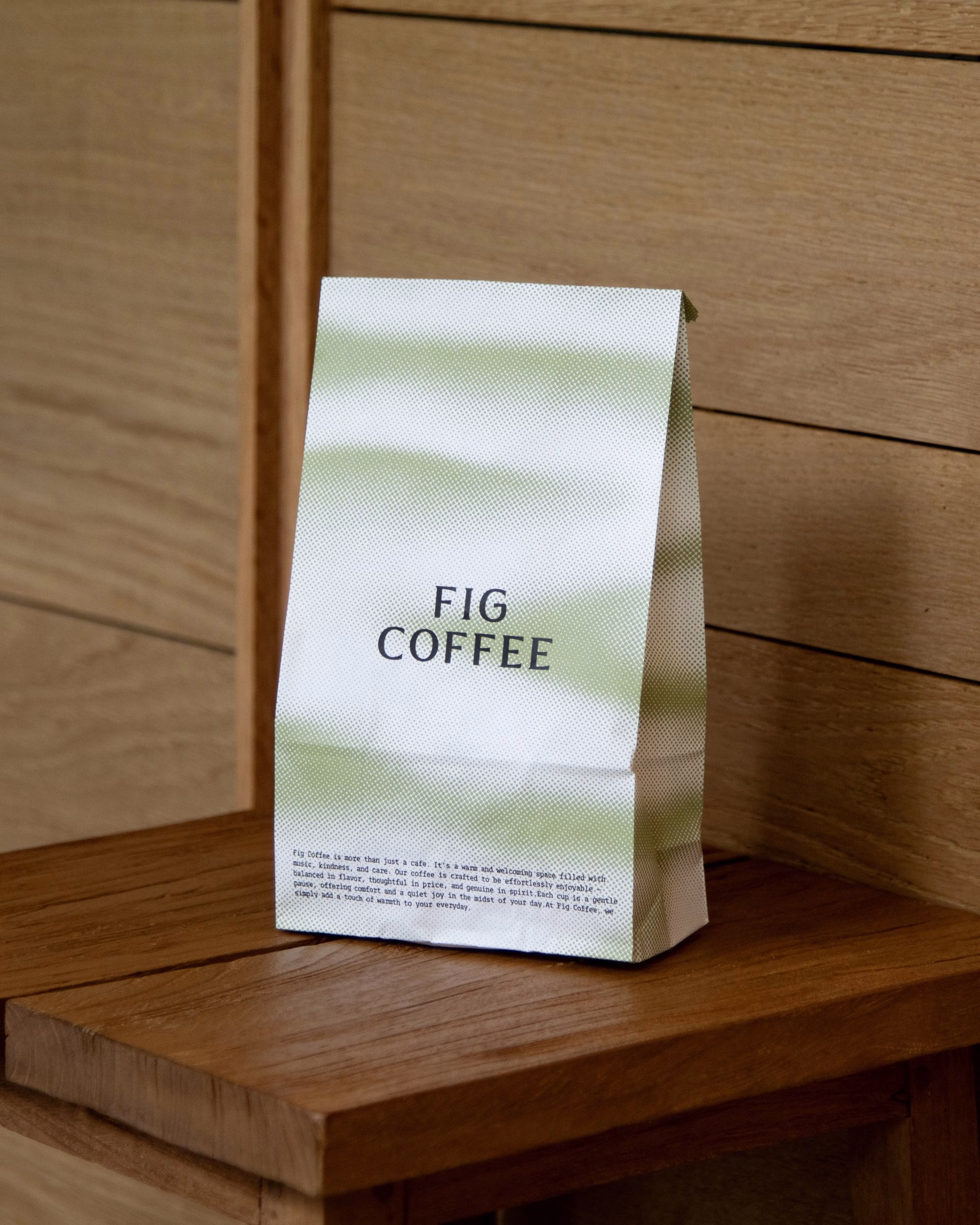 A paper coffee bag with a green gradient design and bold black text reading "FIG COFFEE" rests on a wooden surface.