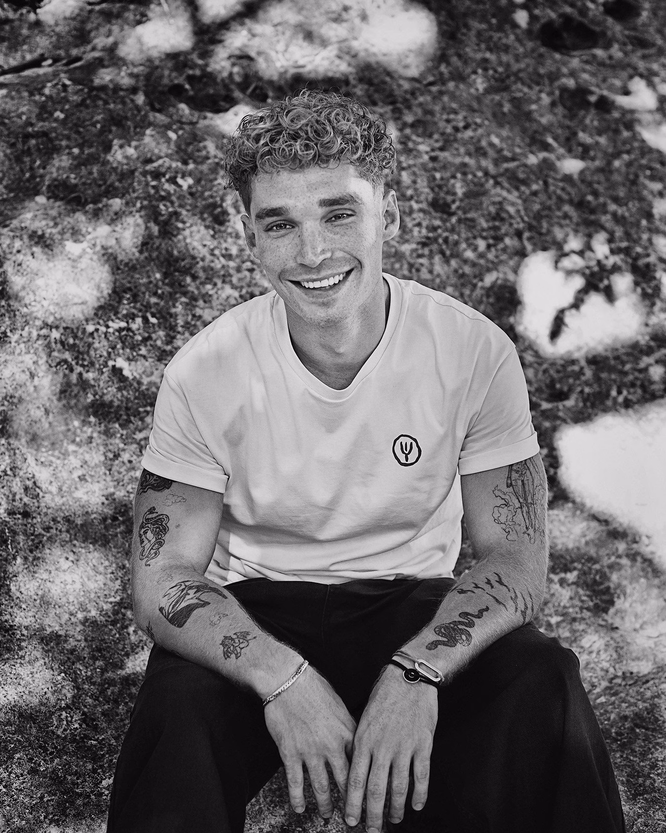 A smiling young man in a white t-shirt with hand-drawn logo sits against a textured rock surface, captured in black and white.