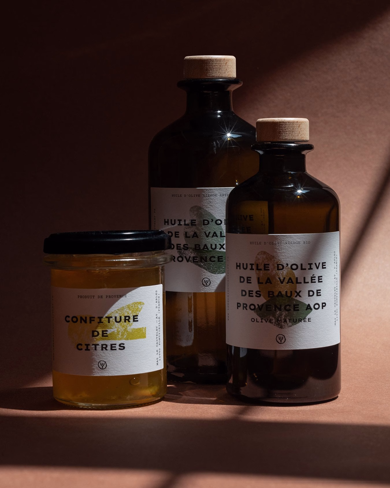 Three jars featuring hand-drawn typography on labels, showcasing olive oil and citrus jam against a warm brown backdrop.