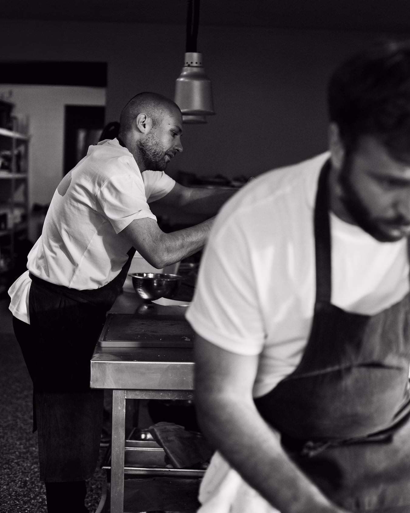 Two chefs in a minimal kitchen, focusing on their tasks, wearing white shirts and dark aprons, captured in black and white.