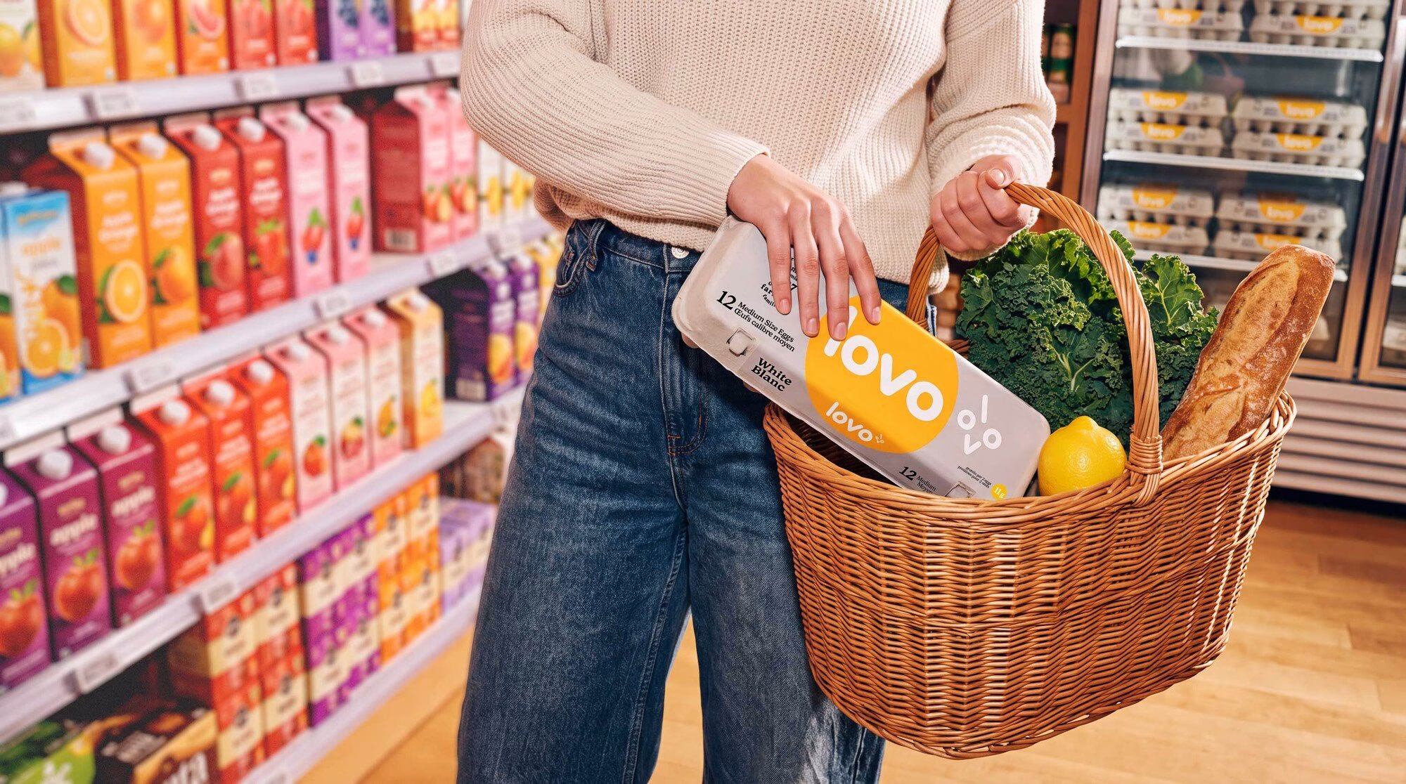 A person holds a wicker basket with LOVO eggs, kale, a lemon, and a baguette in a vibrant grocery aisle.
