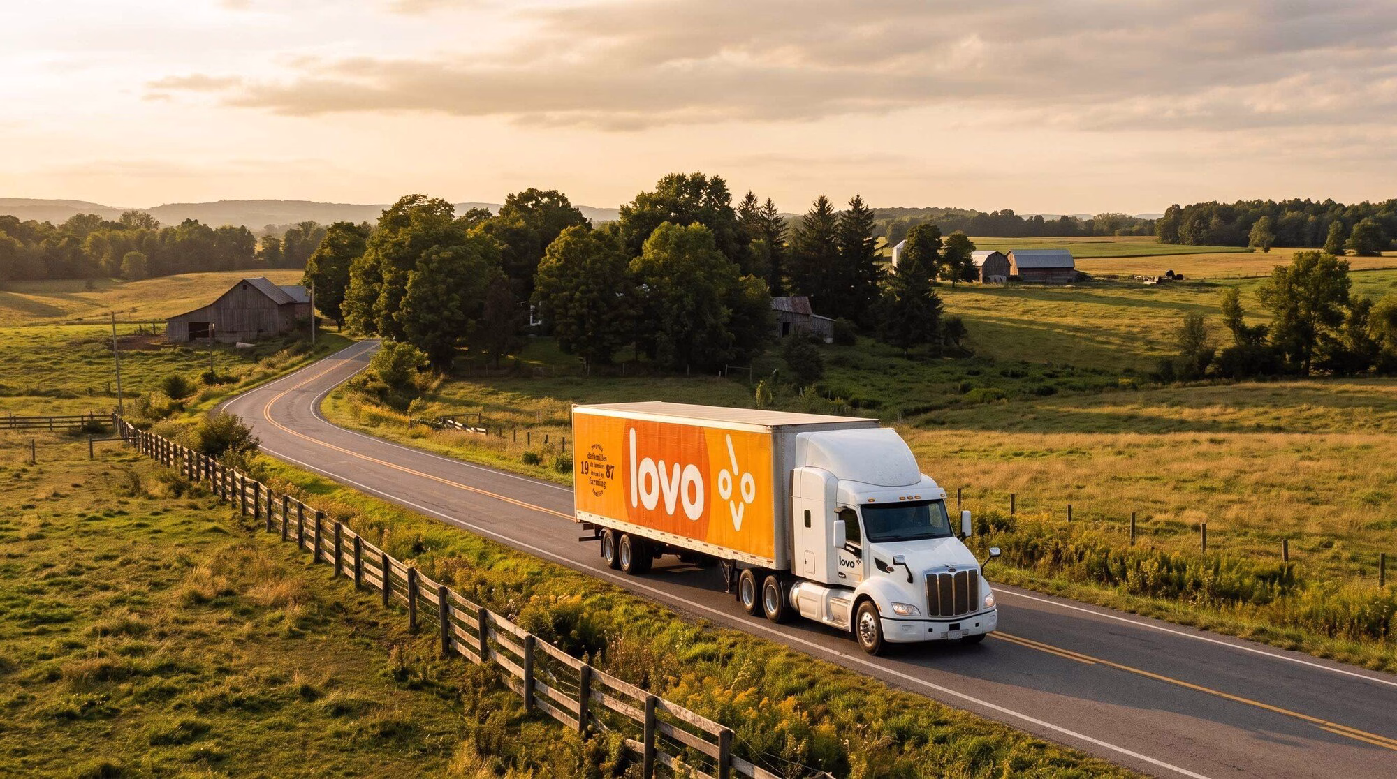 A white truck featuring LOVO's bright orange branding drives along a winding rural road surrounded by green fields and trees.